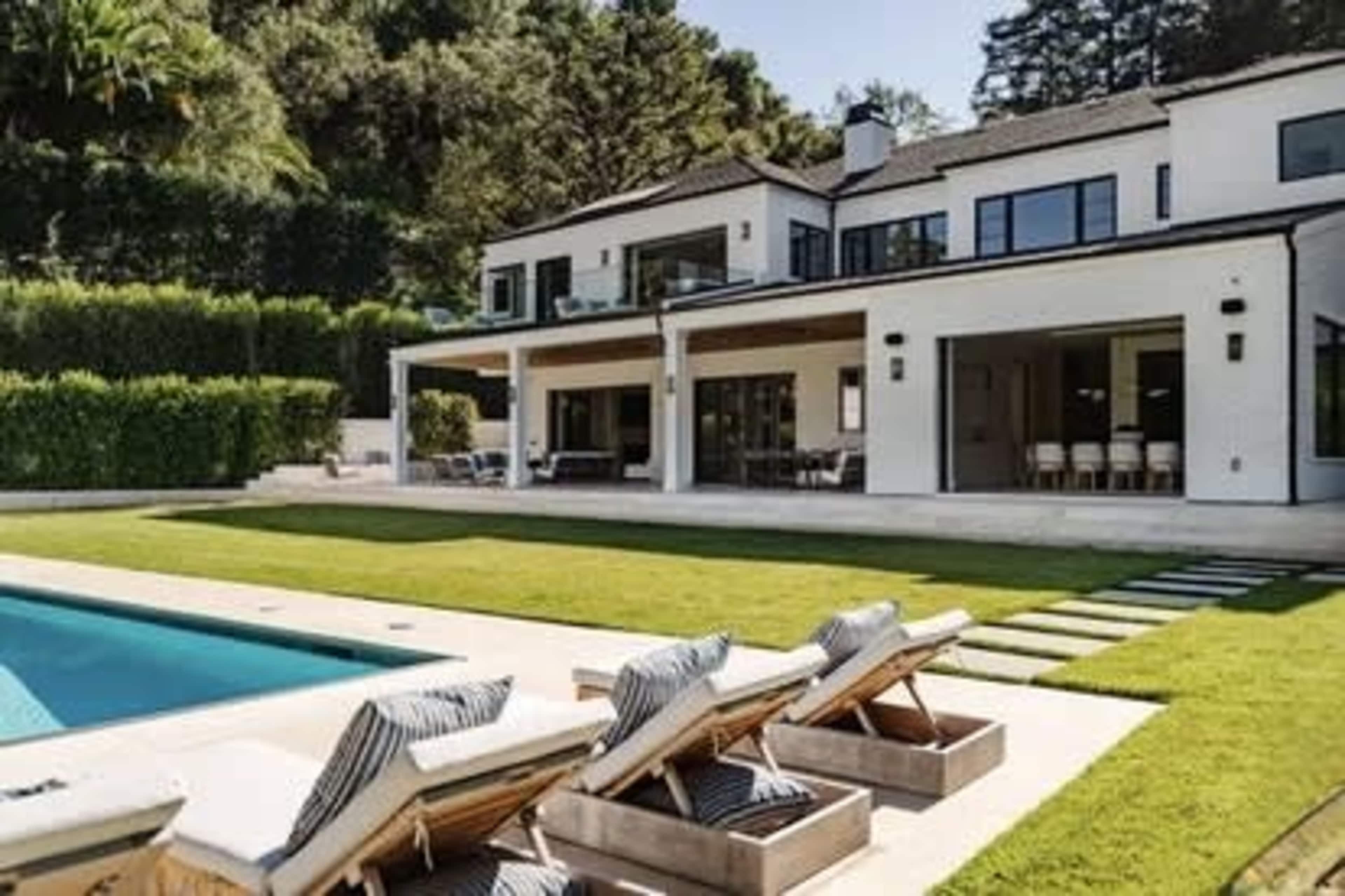 The image shows a modern white villa with a large pool, surrounded by green grass and lounge chairs on a sunny day.