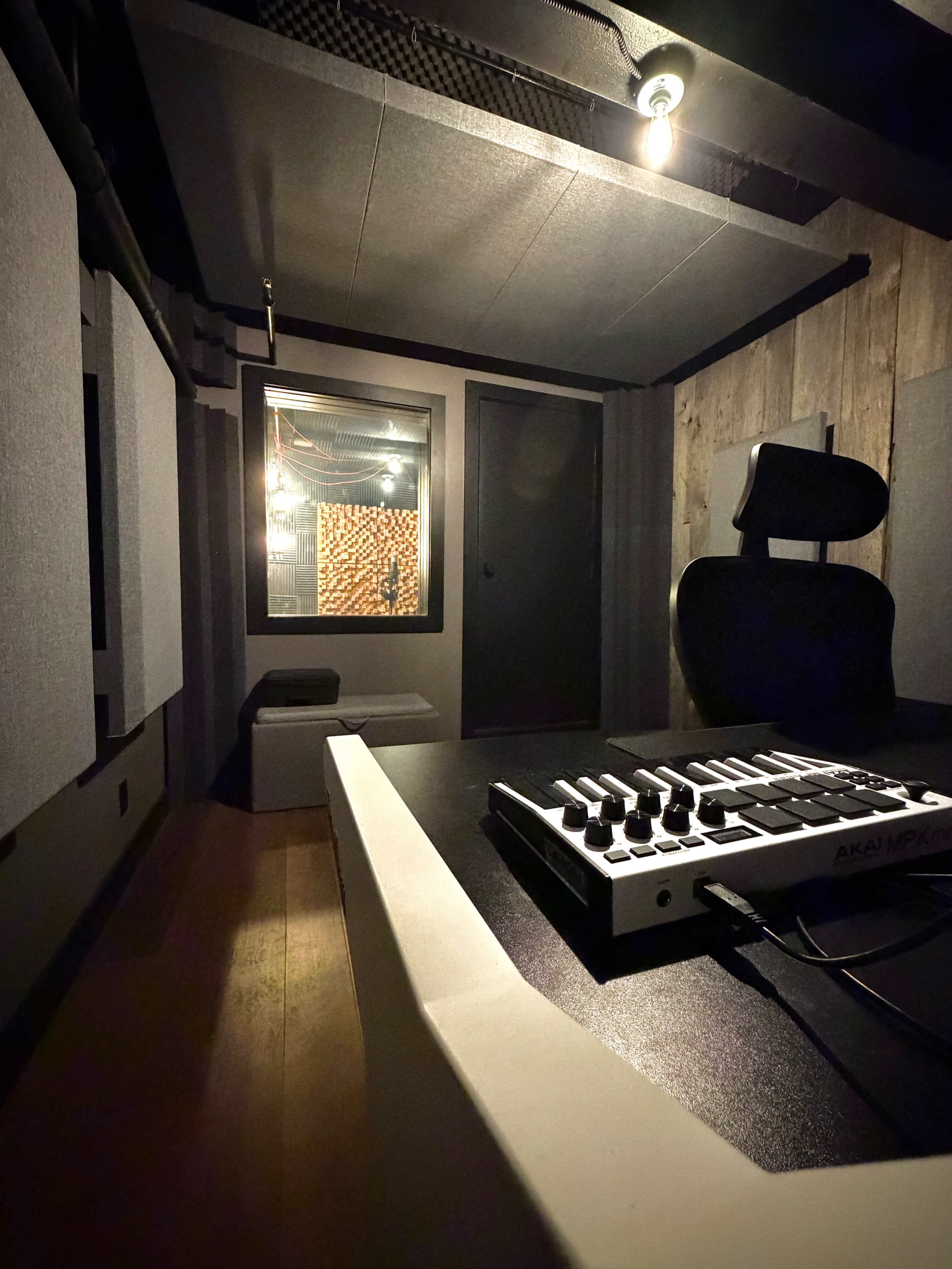 The image shows a sound studio with acoustic panels, a control surface, and a small glass window that leads to another room.