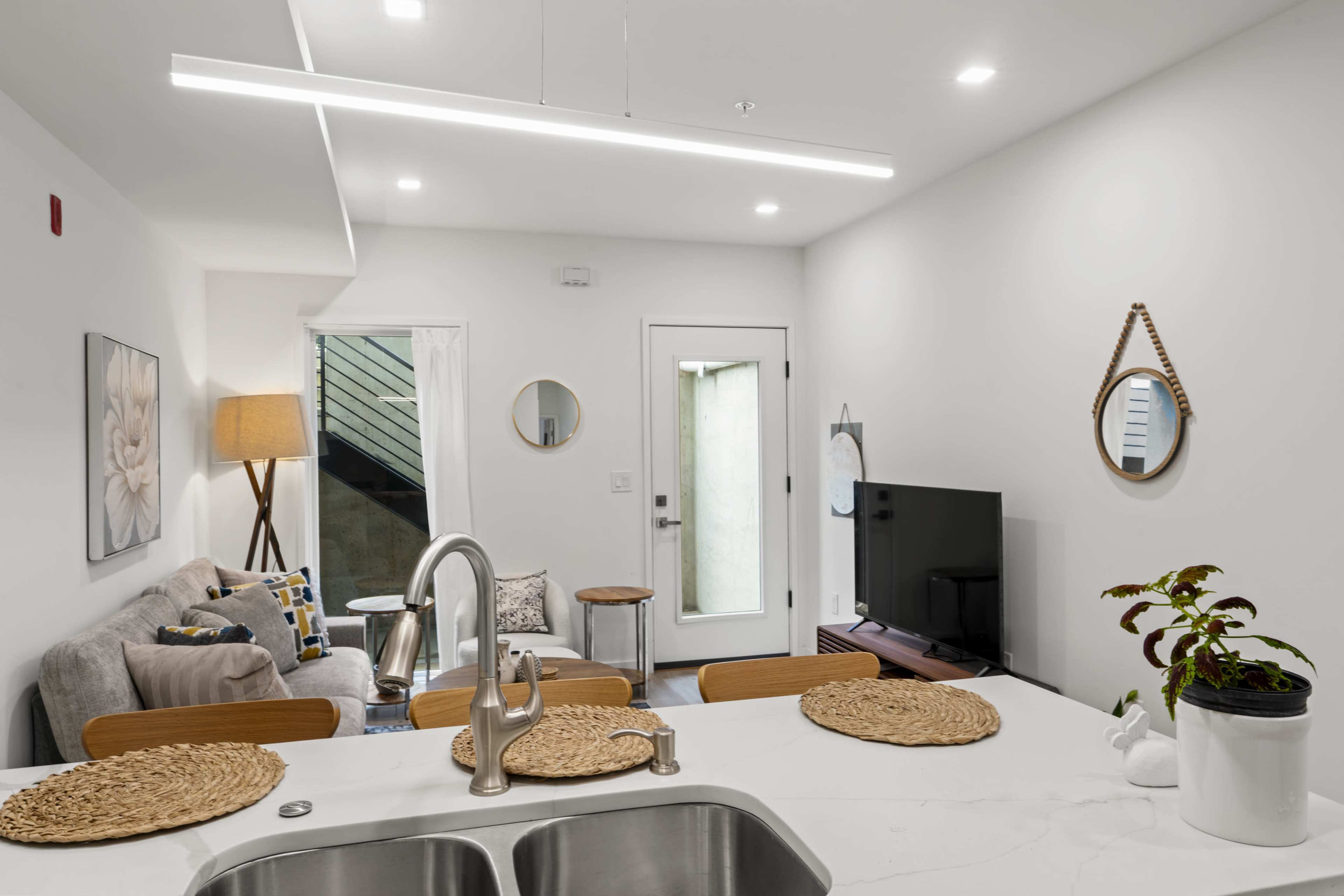 The image shows a modern living room area with a sofa, a television, a coffee table, and a door leading outside, all bathed in natural light.