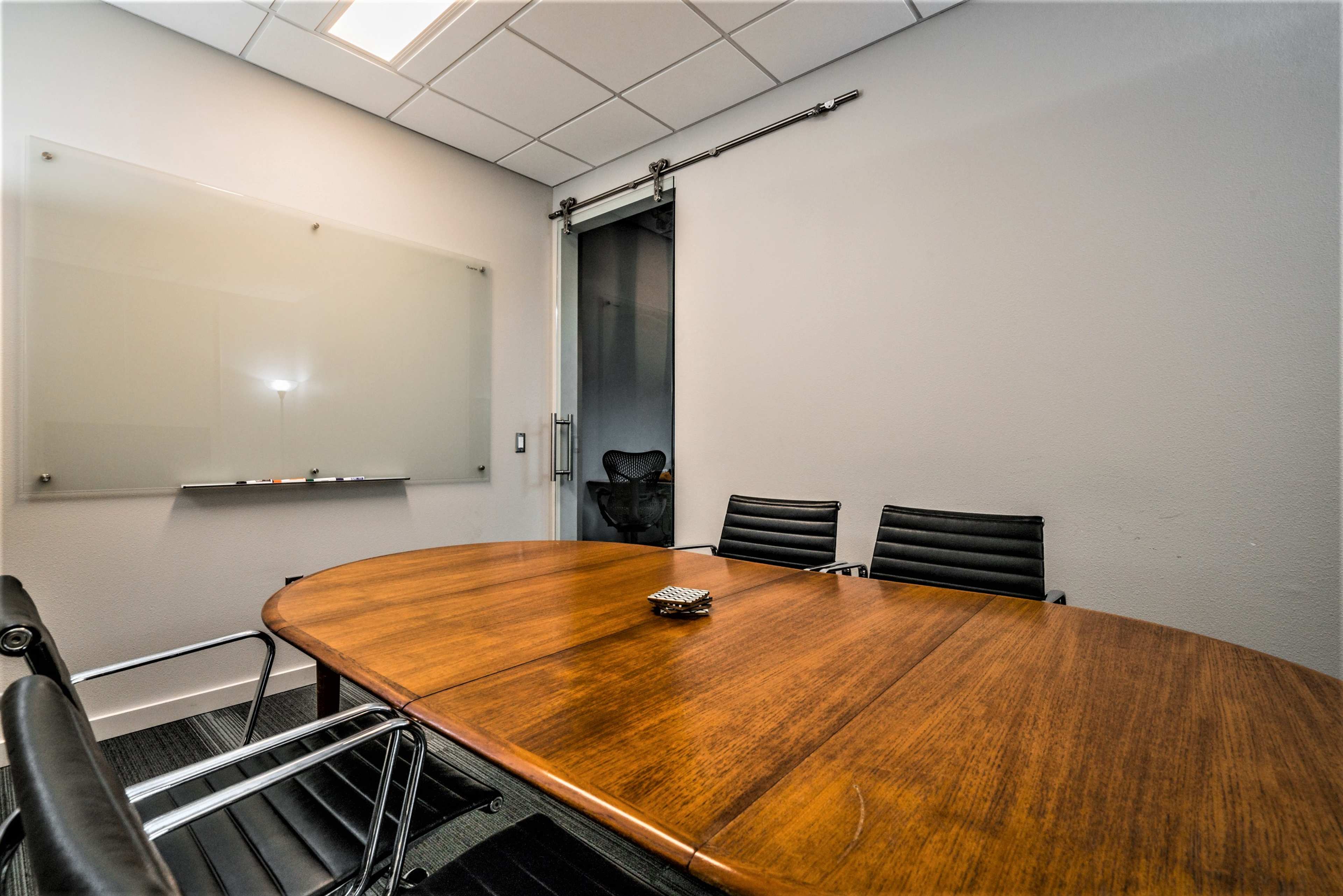 A conference room features a large wooden table surrounded by black leather chairs, with a sliding door leading to another space and a whiteboard on the wall.