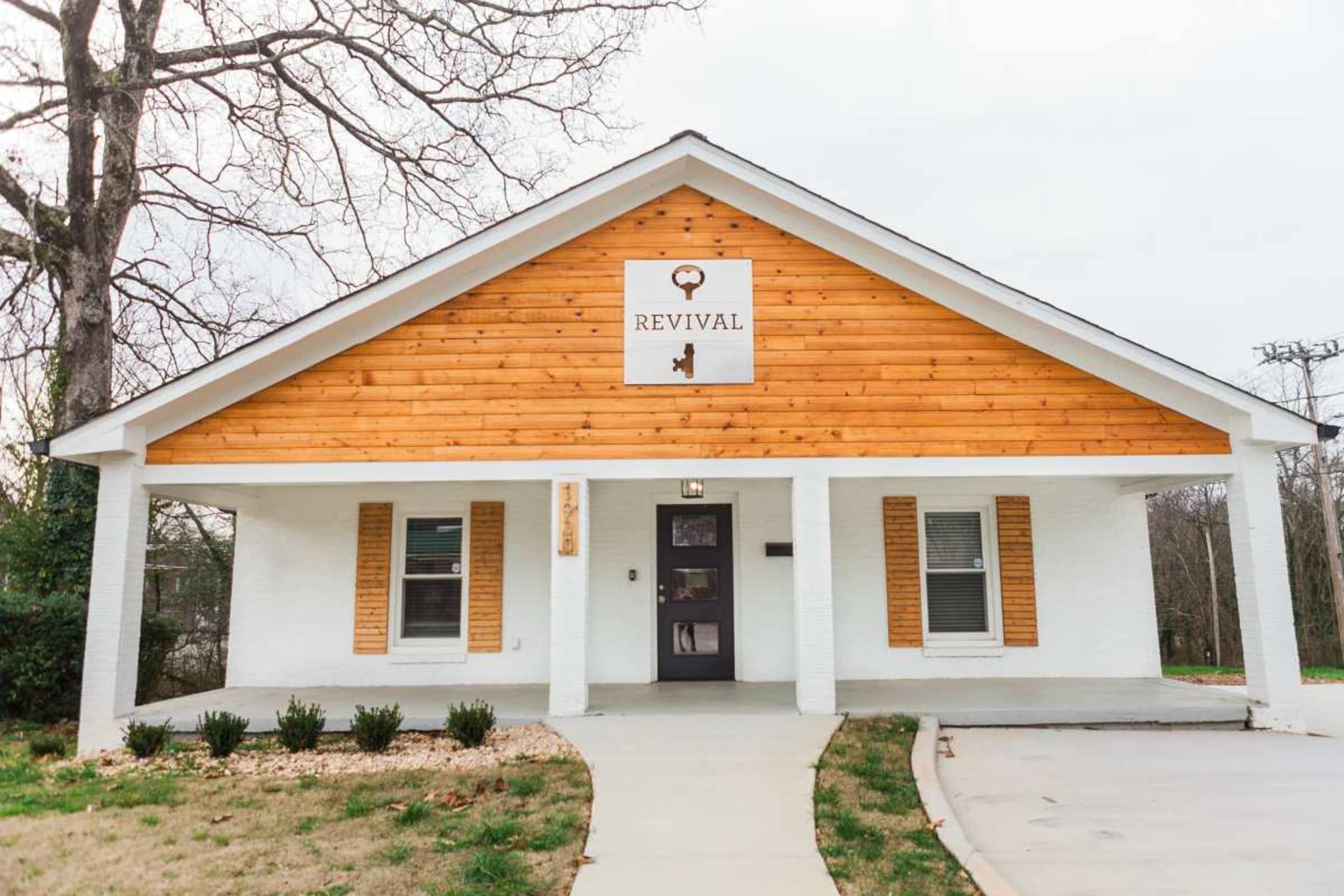 A white and wooden house features a sign labeled "REVIVAL" above the entrance, with a landscaped front yard and a paved walkway.