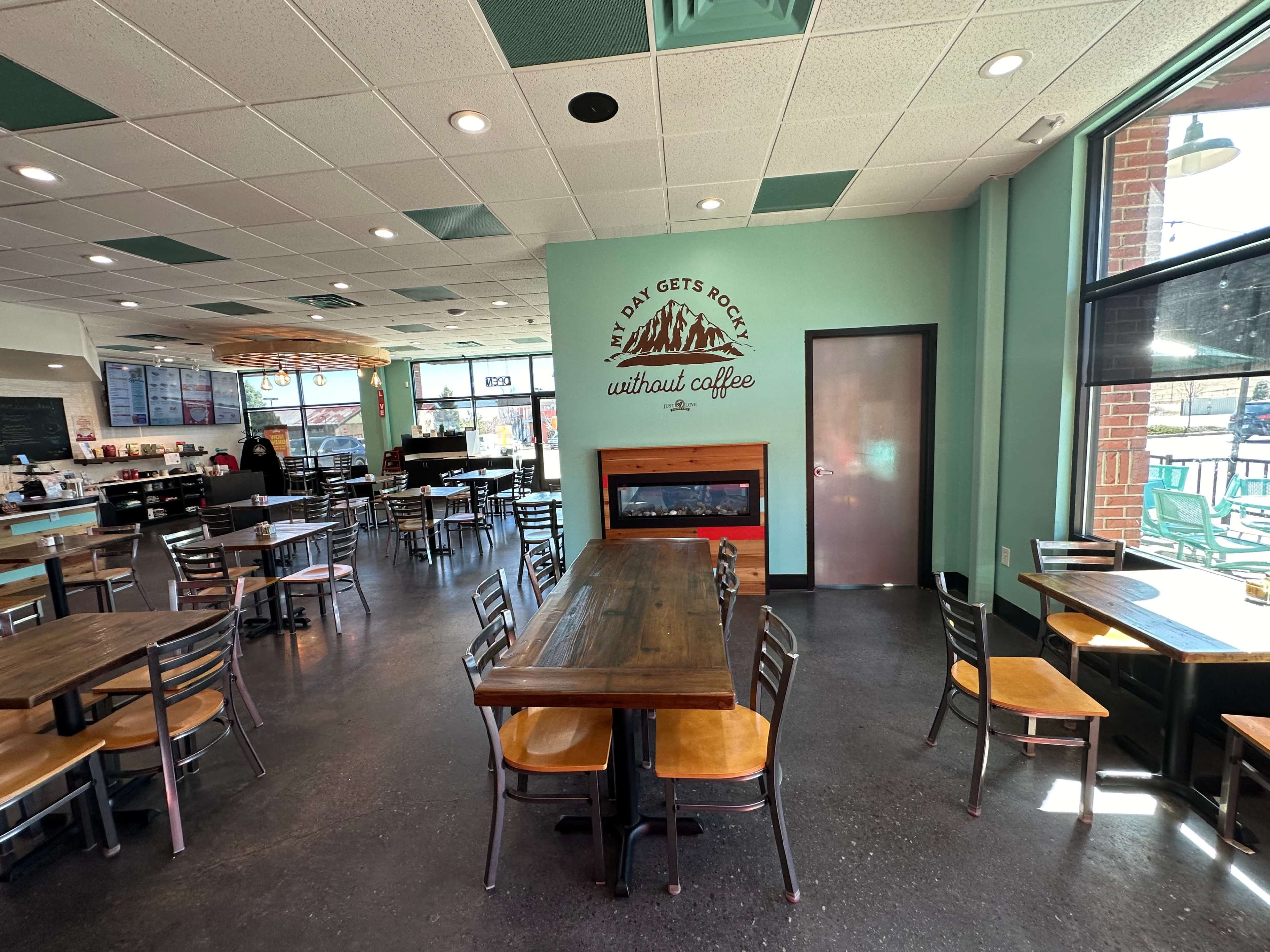 A modern café interior featuring several tables and chairs, with a large window and a wall mural that reads "The Day Gets Rocky Without Coffee."