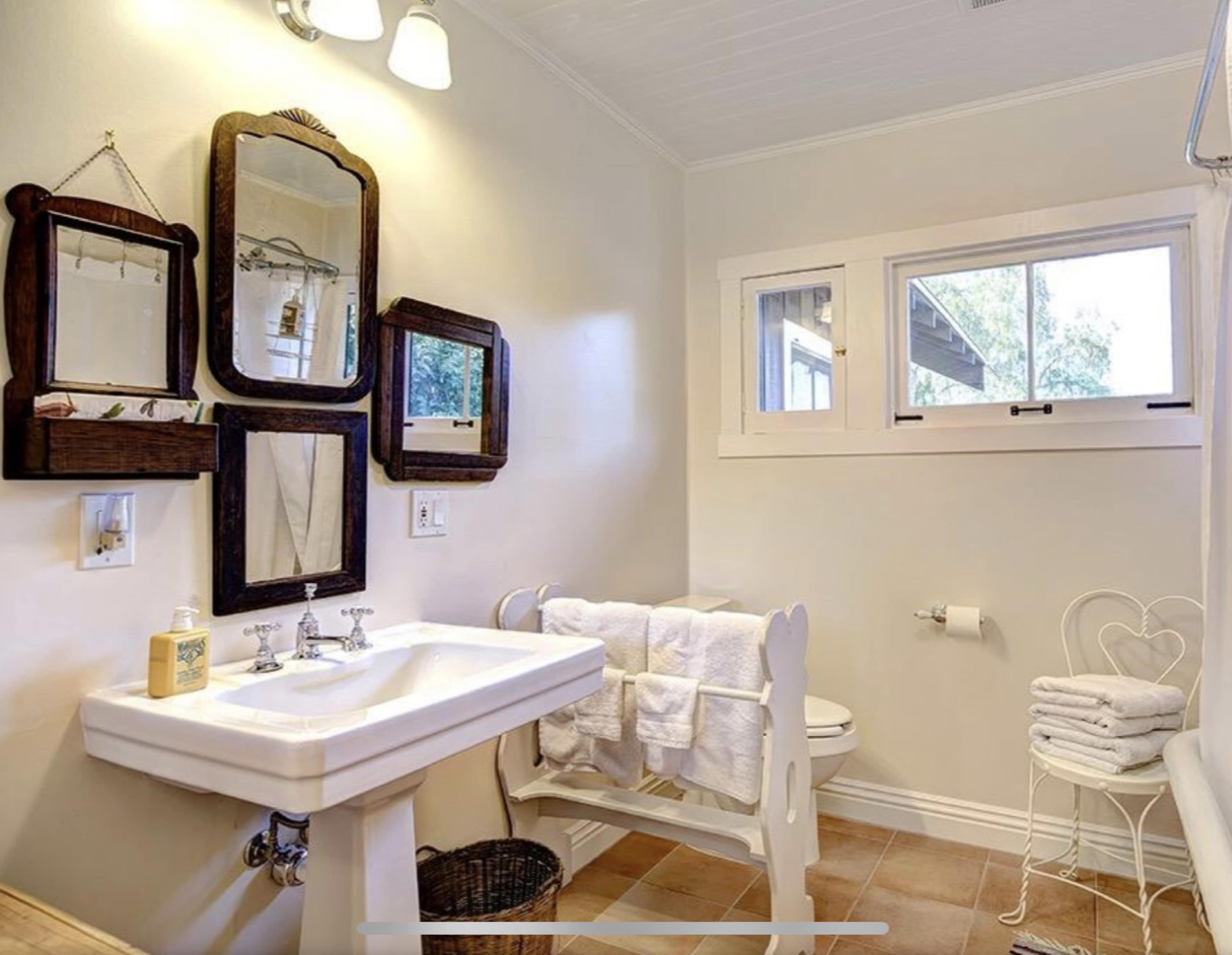 A bathroom with a white sink, three mirrors on the wall, and a window, featuring a towel rack with white towels and a wicker basket on the floor.