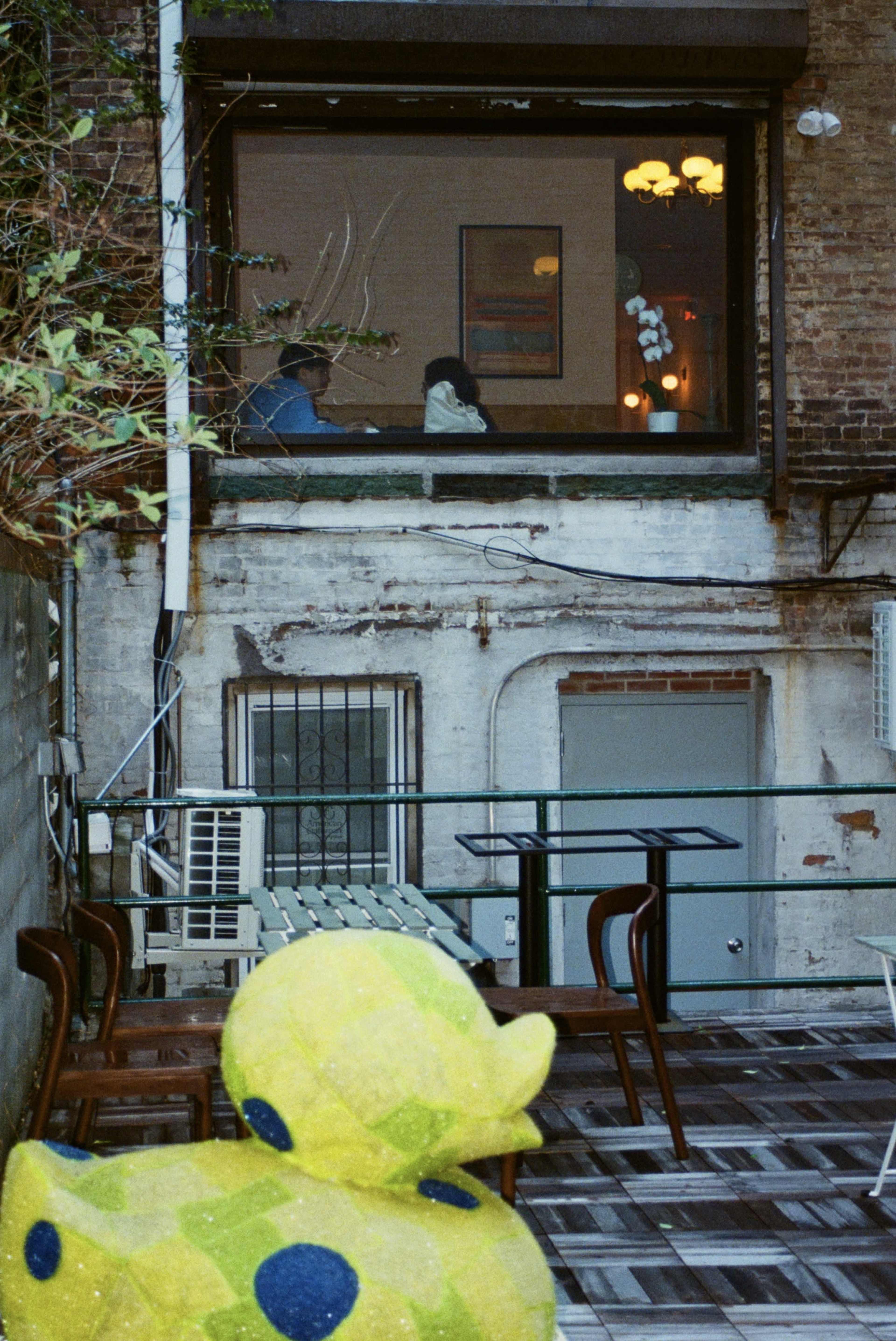 A bright yellow rubber duck sculpture sits in the foreground of a courtyard, overlooking a window where two people are seated inside a room.