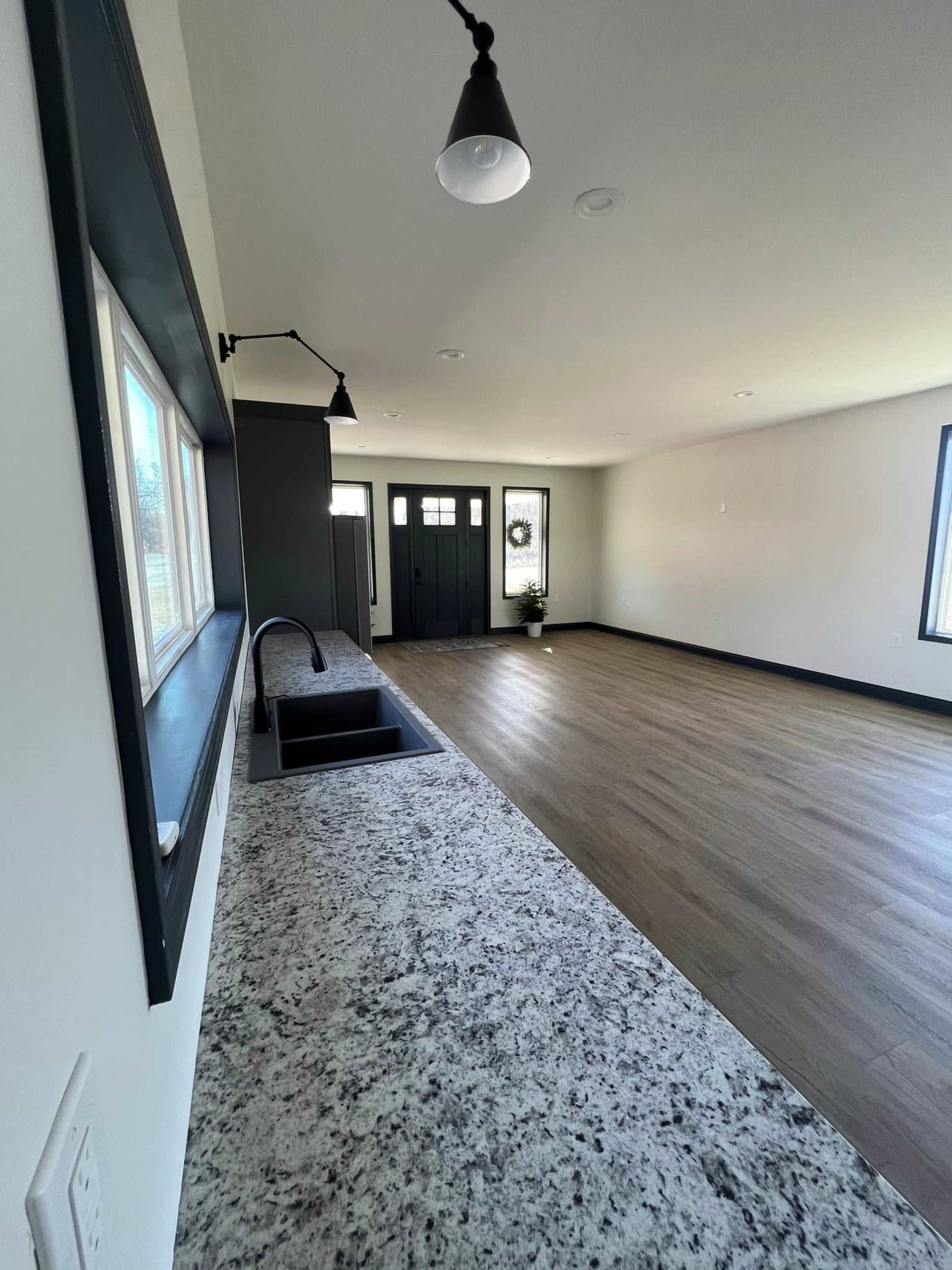 The image shows a spacious, modern interior room featuring a granite countertop, a black sink, and a dark front door in the background.