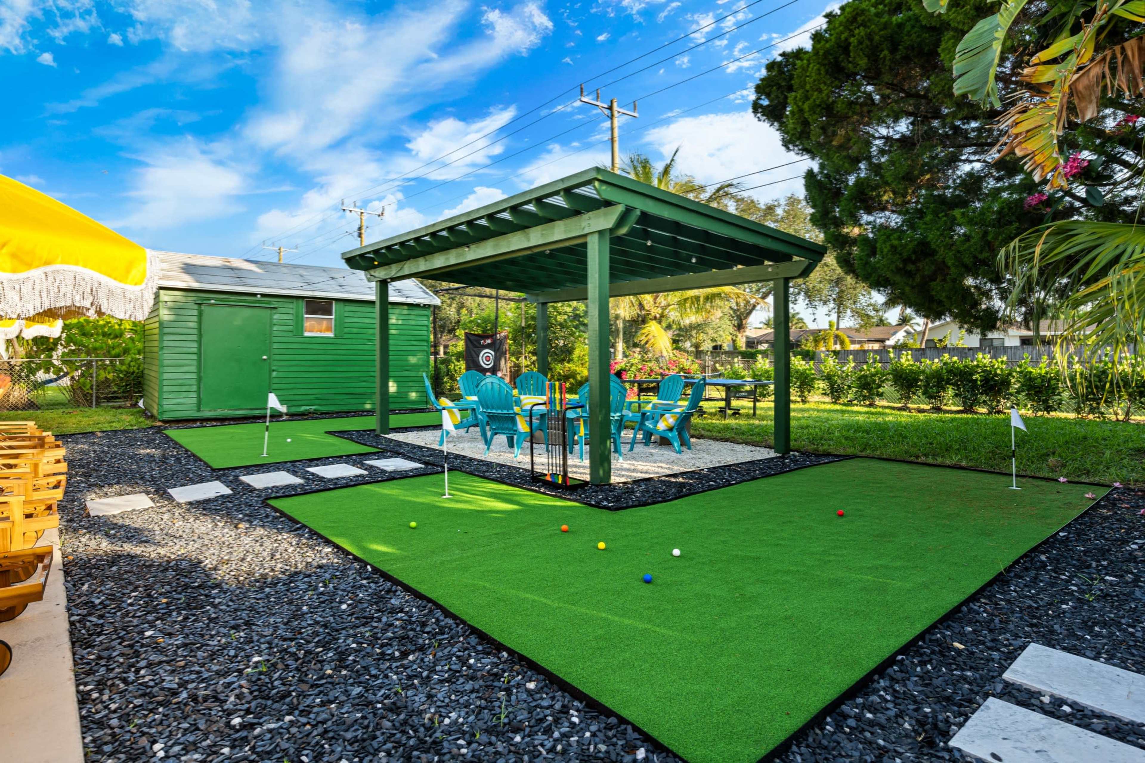 The image shows a small green putting green area surrounded by artificial turf and gravel, with a covered seating area and colorful chairs nearby.