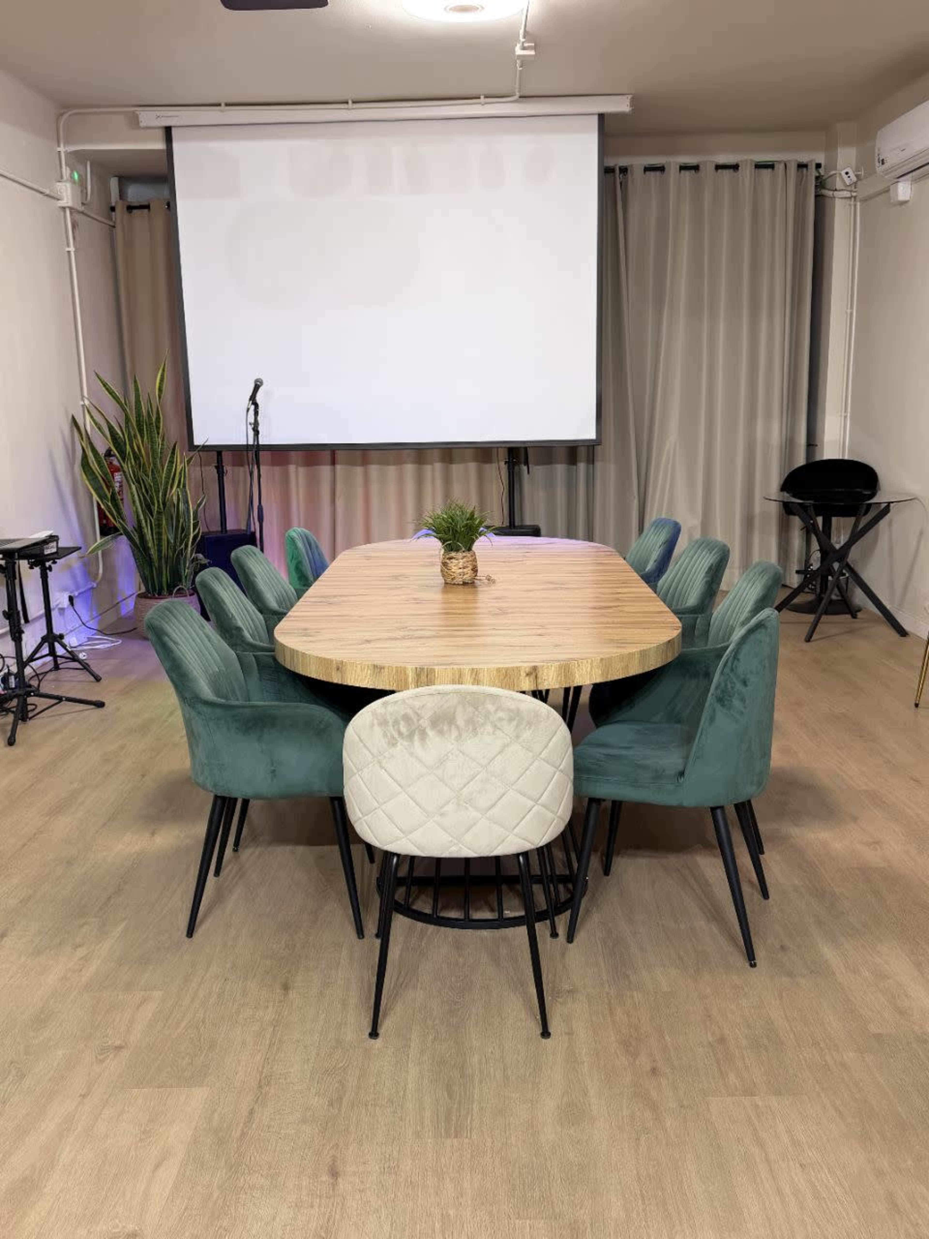 The image shows a round wooden table surrounded by eight upholstered chairs in a room with a projector screen and plant decor.