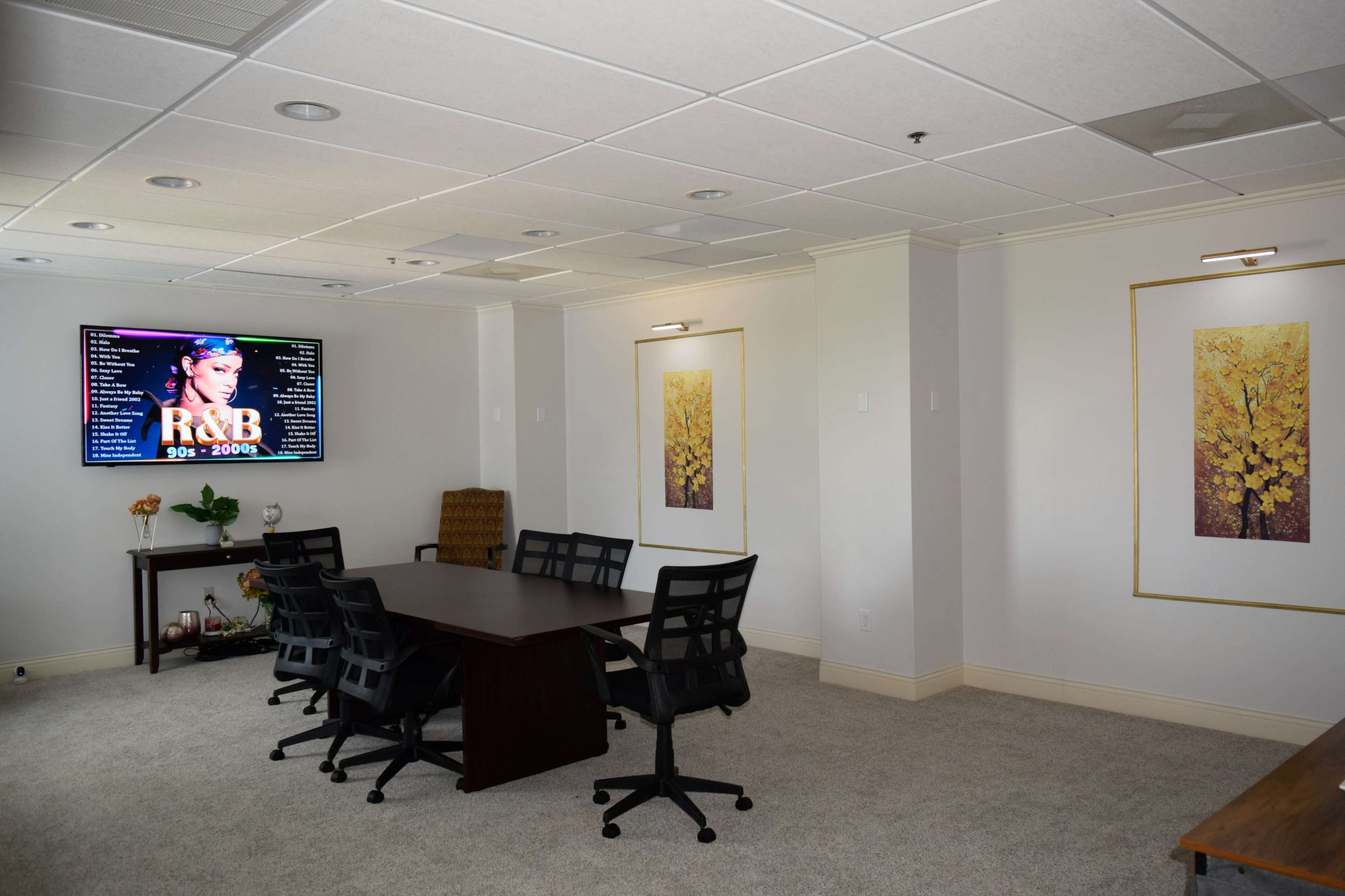 A conference room features a large screen displaying R&B music, surrounded by two framed leaf prints and a table with six chairs.