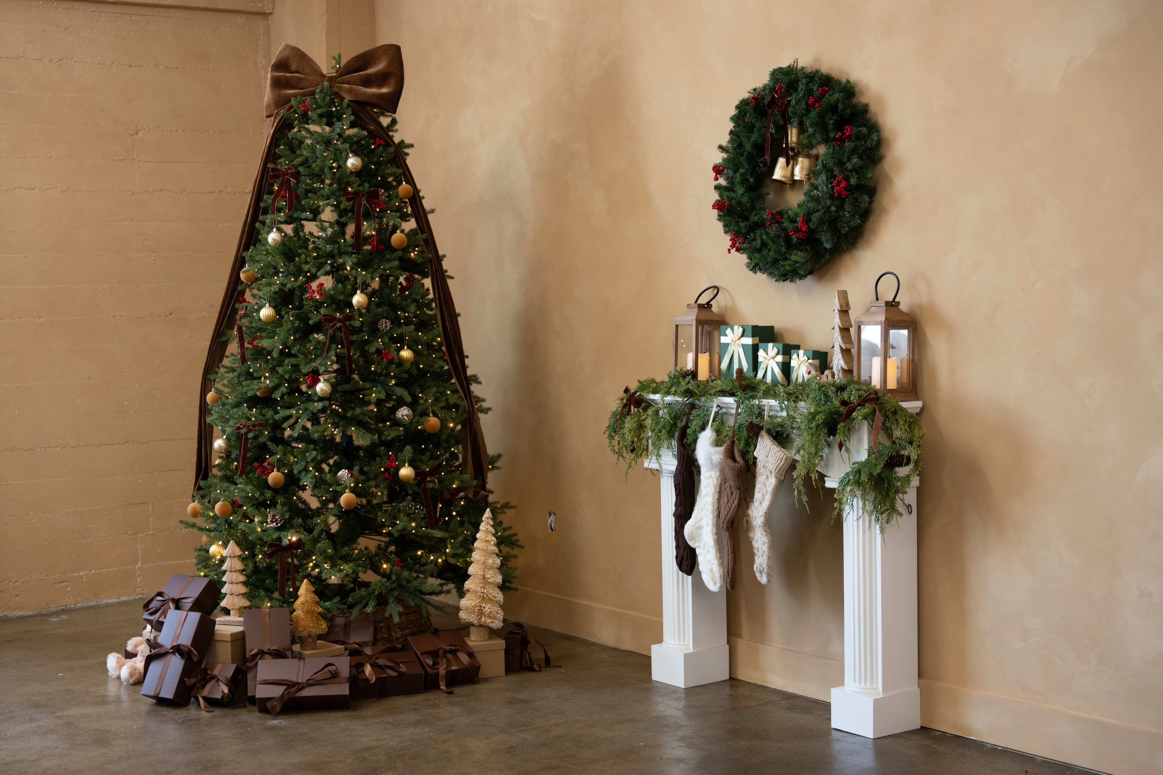 A decorated Christmas tree is placed next to a white mantel adorned with stockings, greenery, and lanterns, against a tan wall.