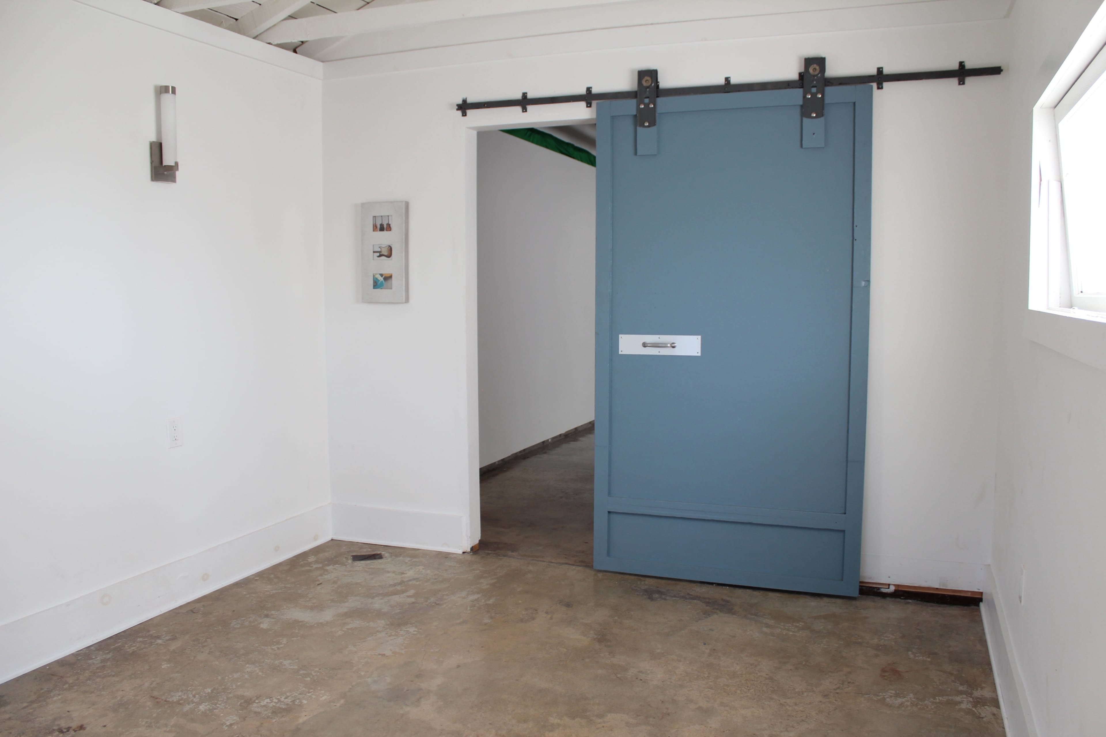The image shows a room with a concrete floor, a blue sliding barn door, and white walls with minimal decor.