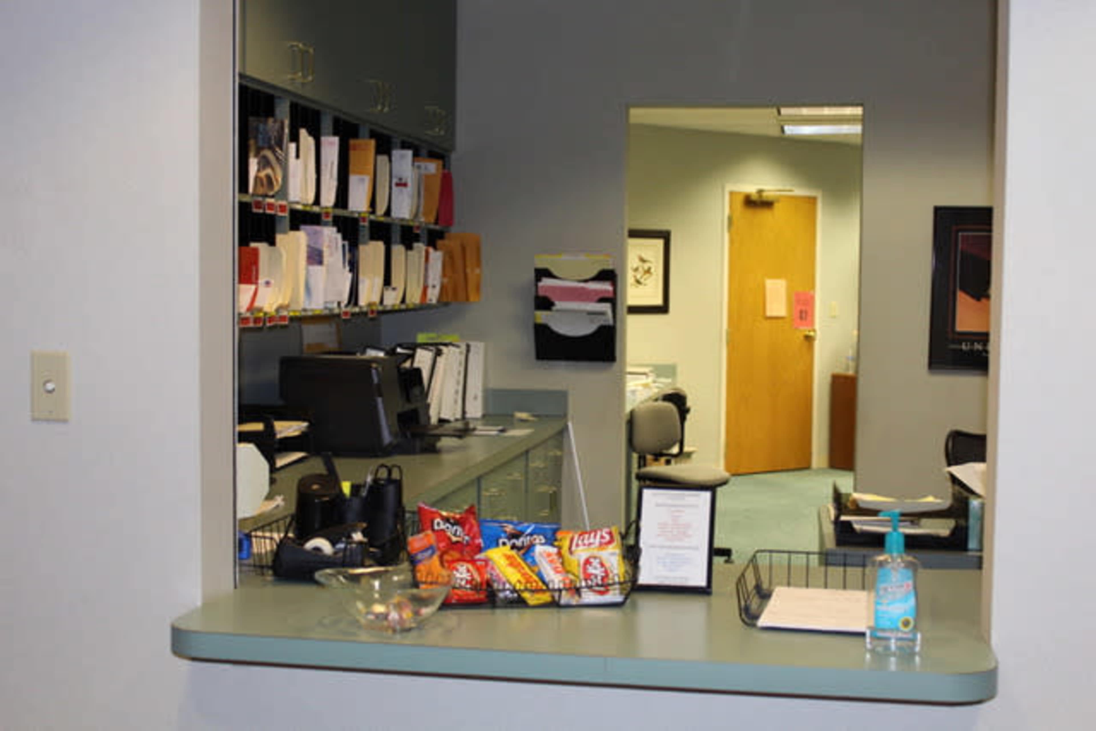 An office reception area features a counter with snacks and a hand sanitizer, while a hallway leads to an office space in the background.