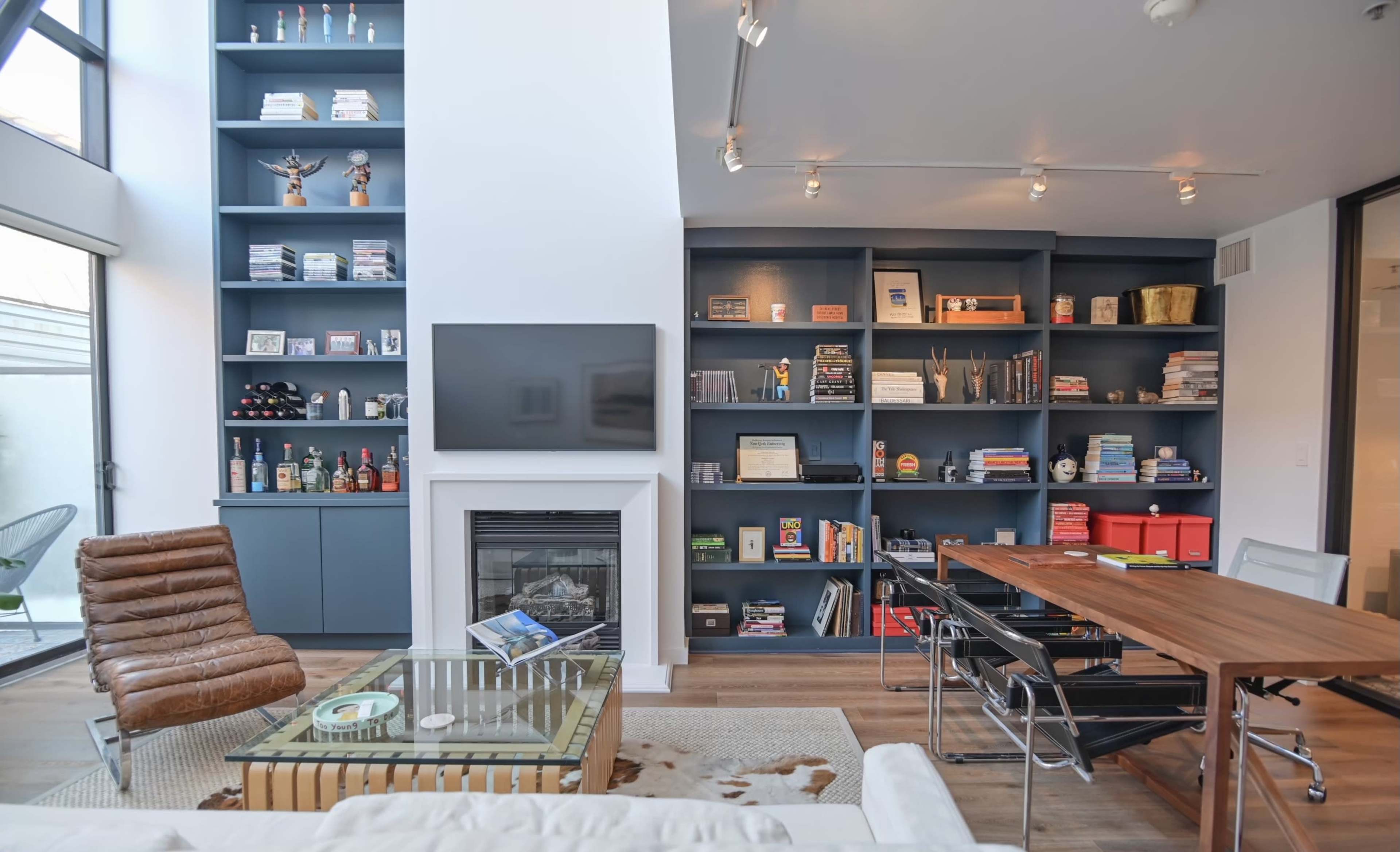 The image shows a modern living room featuring a blue shelving unit filled with books and decorative items, a brown leather chair, a glass coffee table, and a wooden dining table.