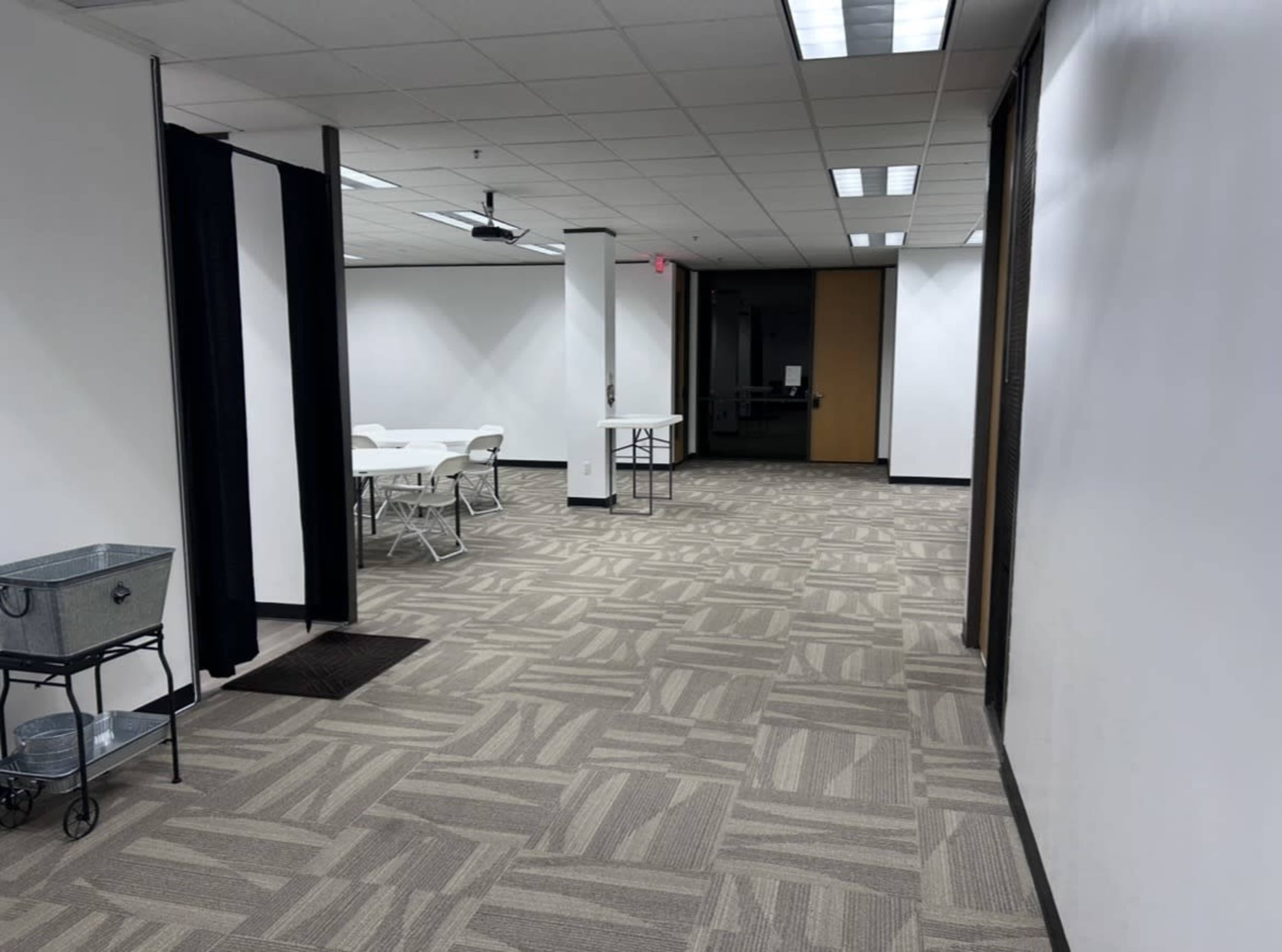 The image shows a spacious, well-lit corridor with a patterned carpet, leading to a room with tables and chairs set up for use.