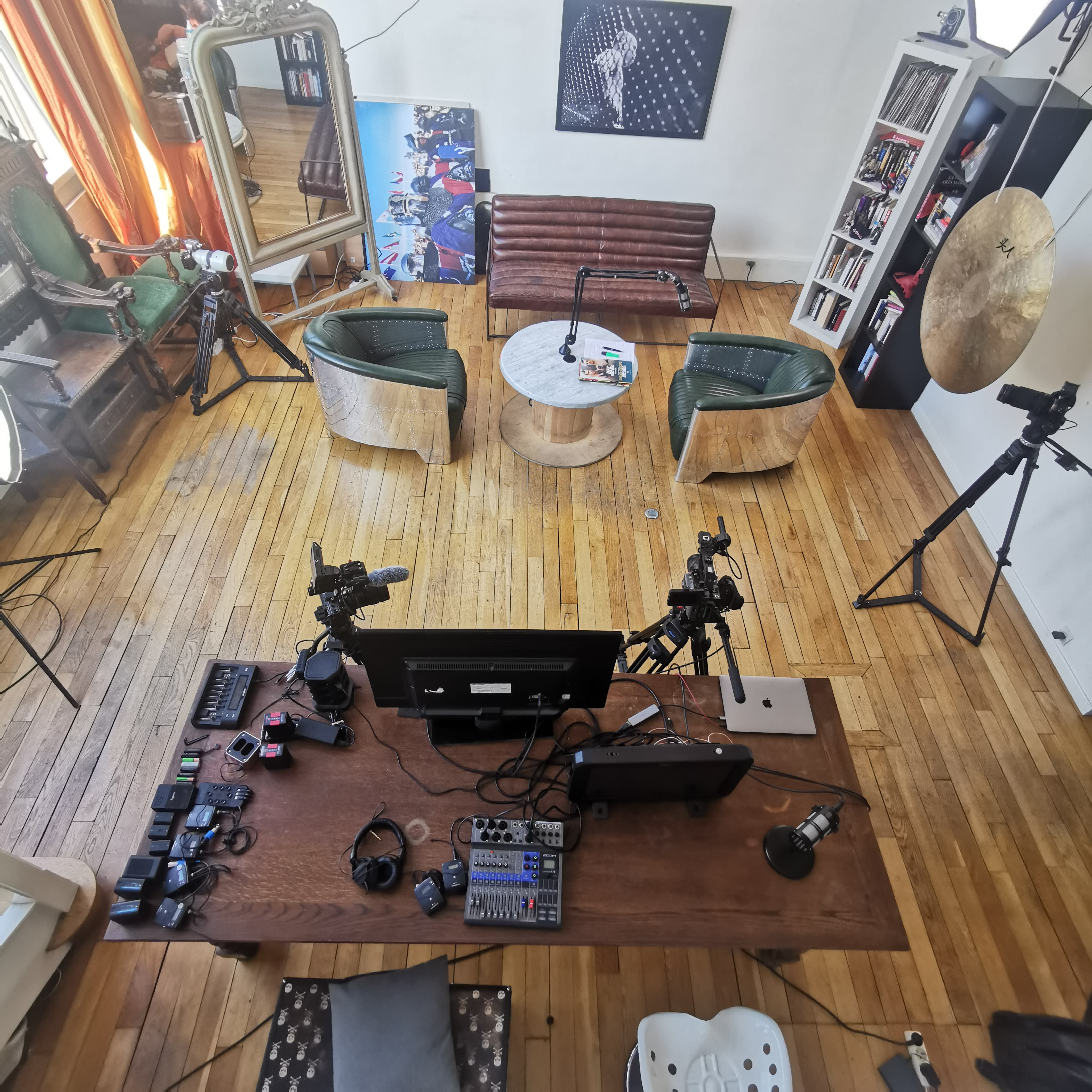 A modern studio setup with a wooden table, two green chairs, a brown couch, multiple cameras, and a large mirror in a brightly lit room.
