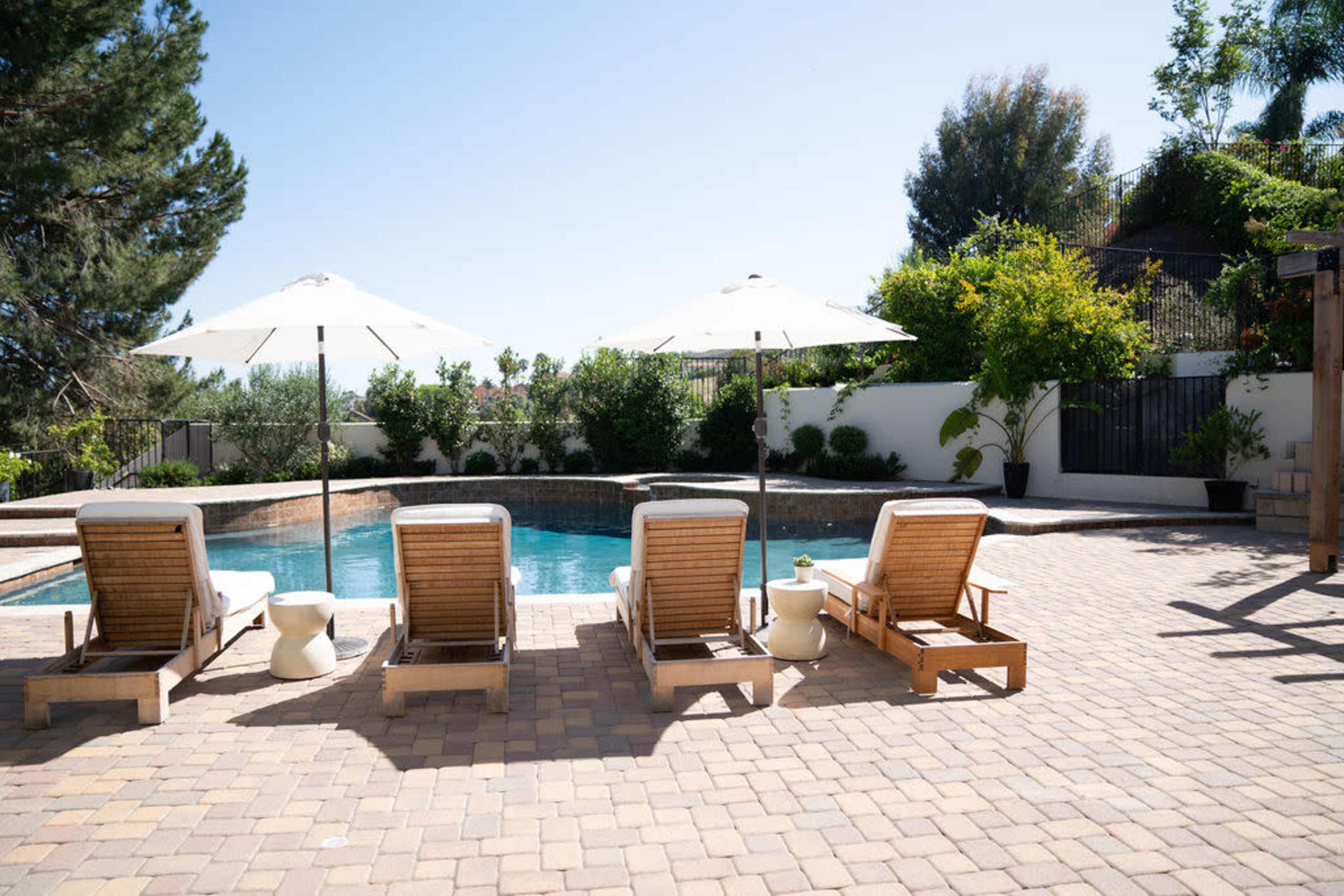 A poolside area with four wooden lounge chairs under white umbrellas, surrounded by greenery and a stone patio.