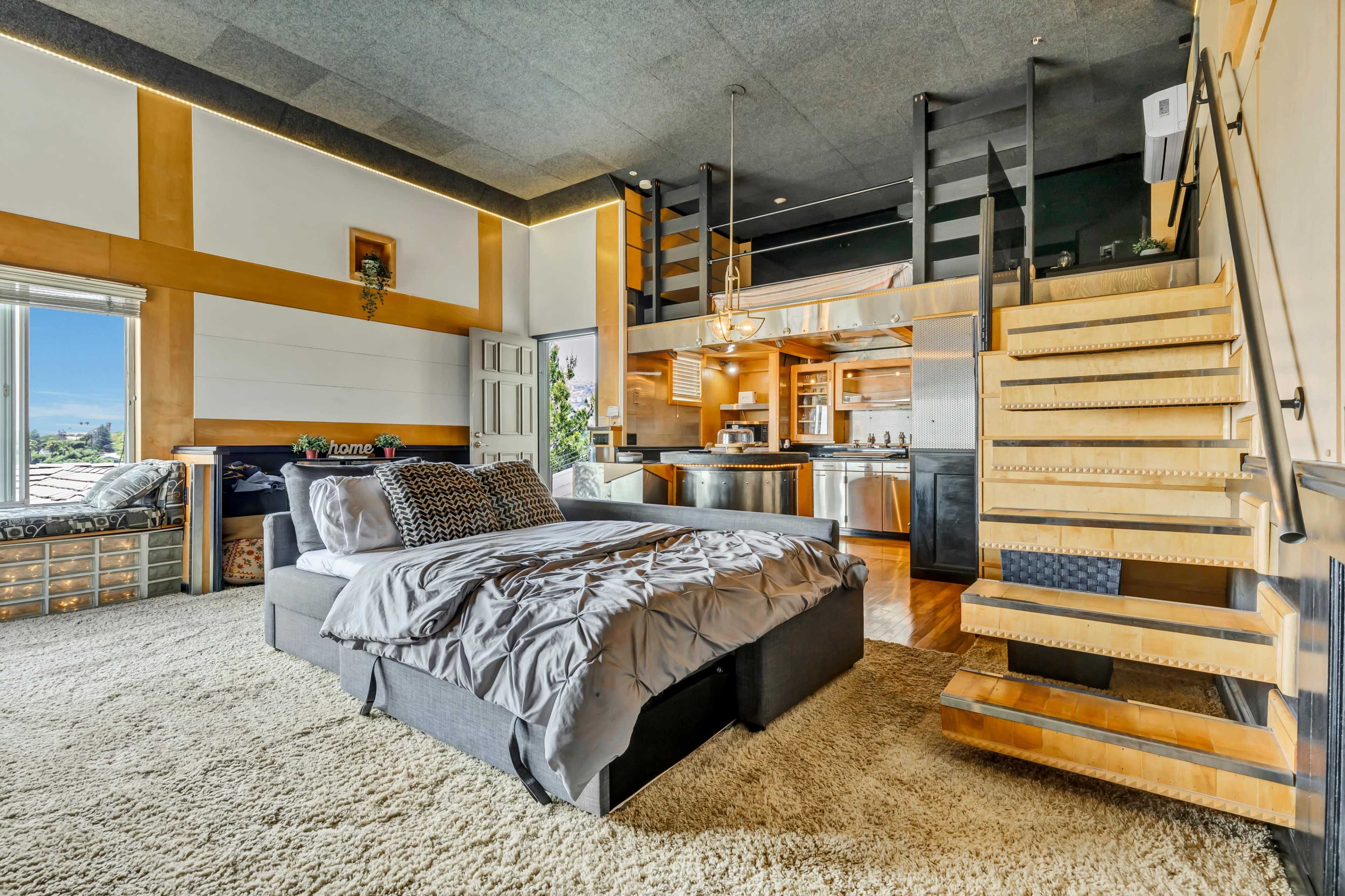 A modern loft-style bedroom with a bed on a plush rug, a wooden staircase leading to a lofted area, and an open kitchen in the background.