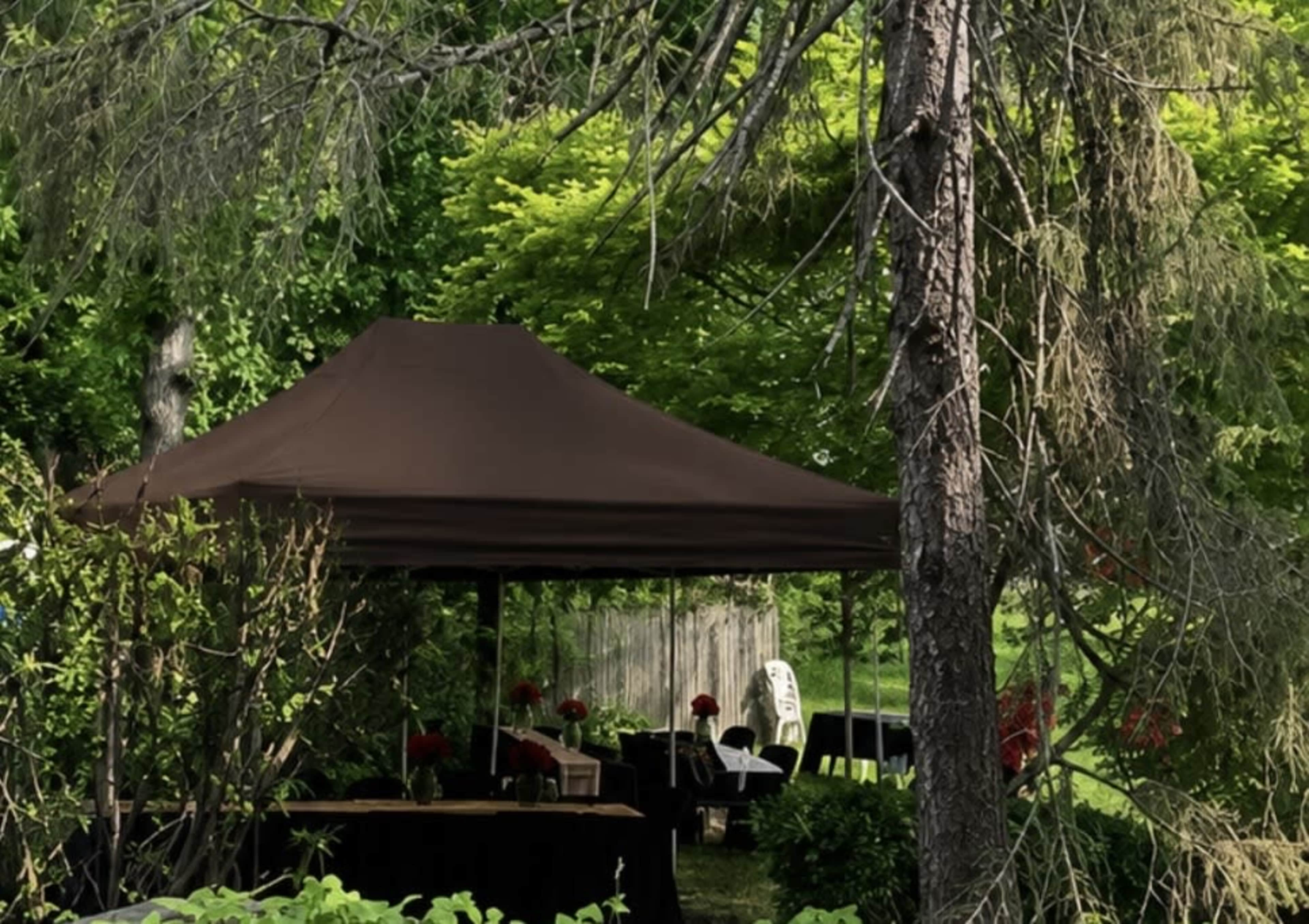 A brown gazebo is set up among trees, with tables adorned with red flowers in the background.