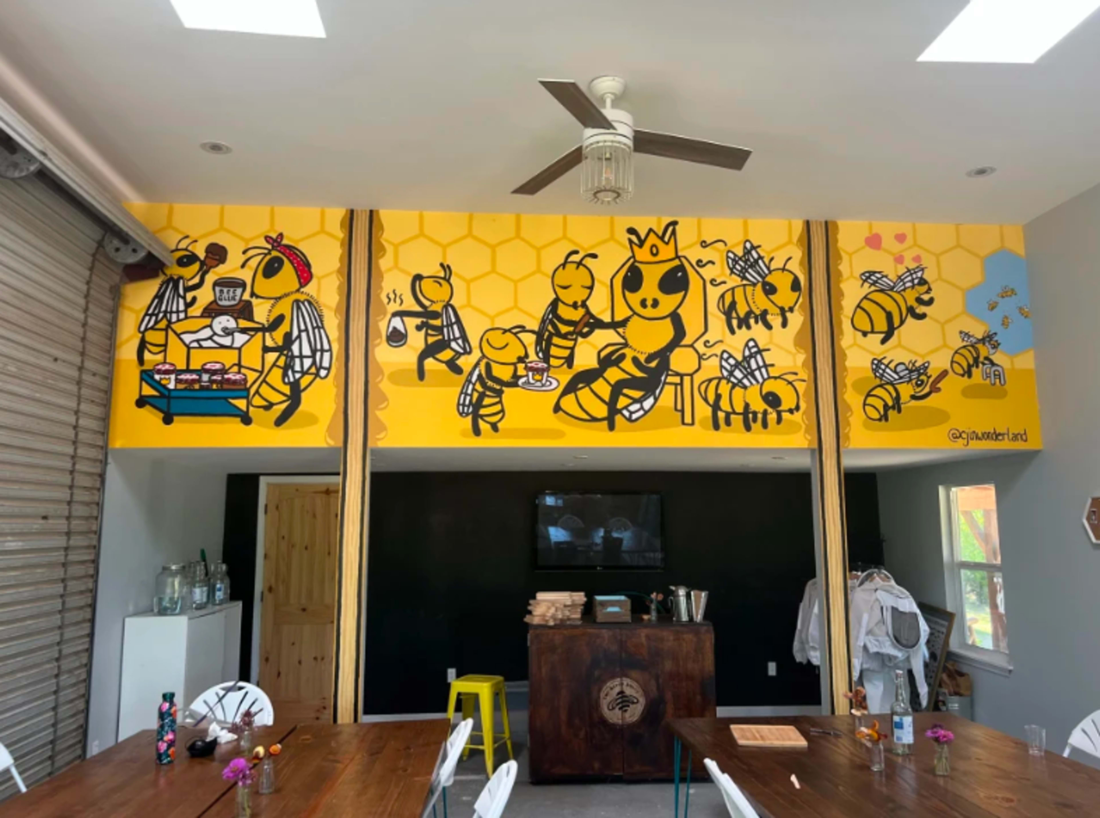 The image shows a colorful mural of cartoon bees in a hive setting on the wall of a room with a table and chairs.