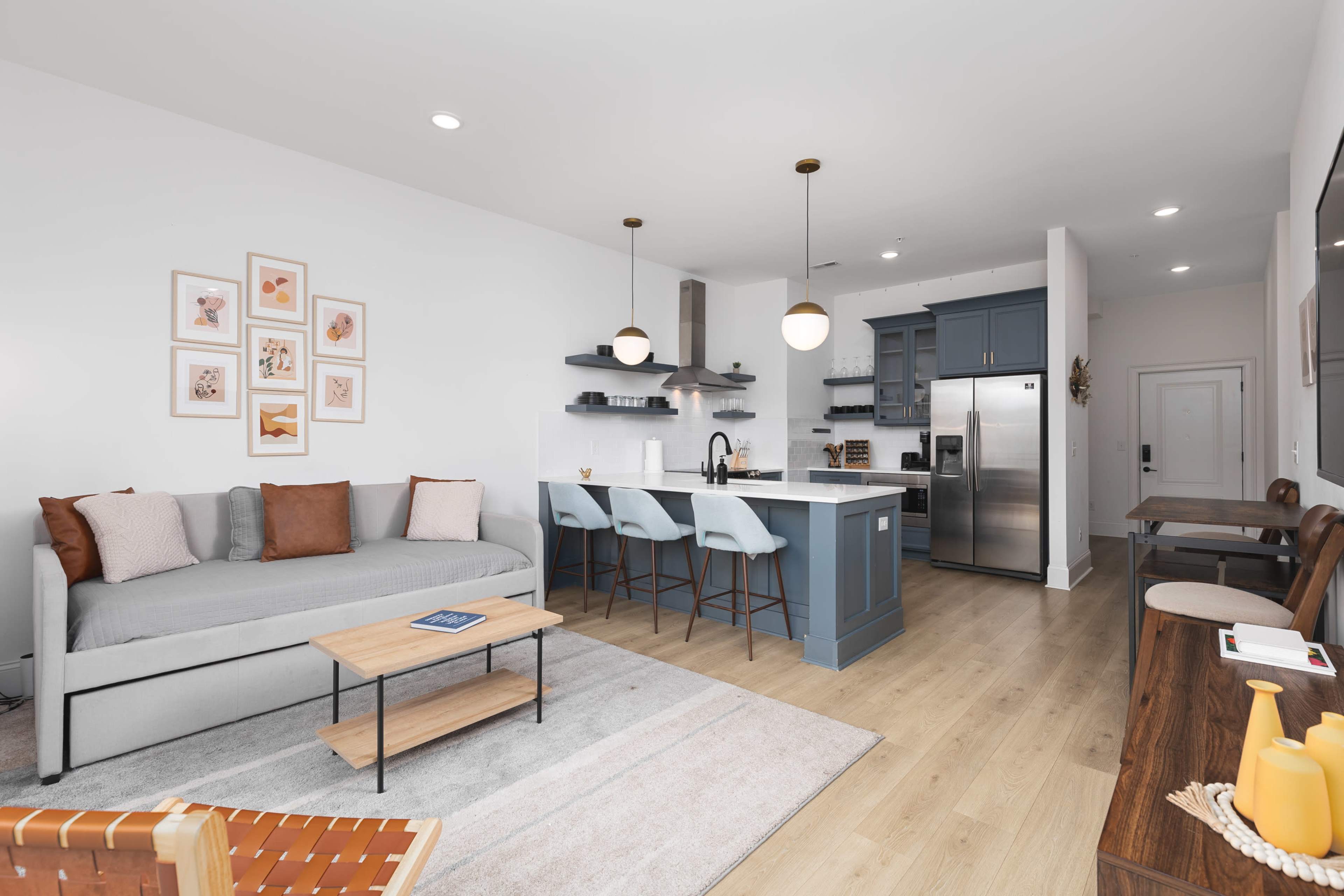 The image depicts a modern open-concept living space featuring a gray sofa, a wooden coffee table, and a kitchen with blue cabinets and bar stools.