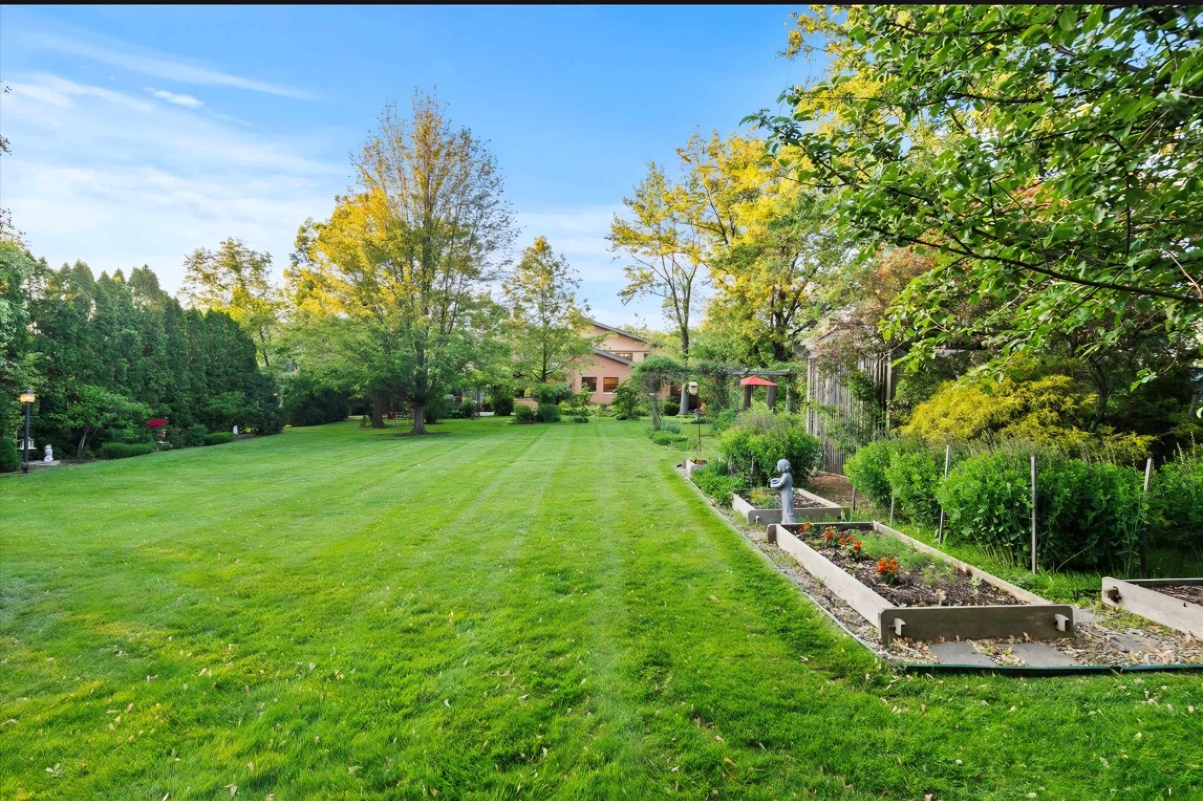 The image shows a spacious green lawn bordered by trees and garden beds, with a house visible in the background.
