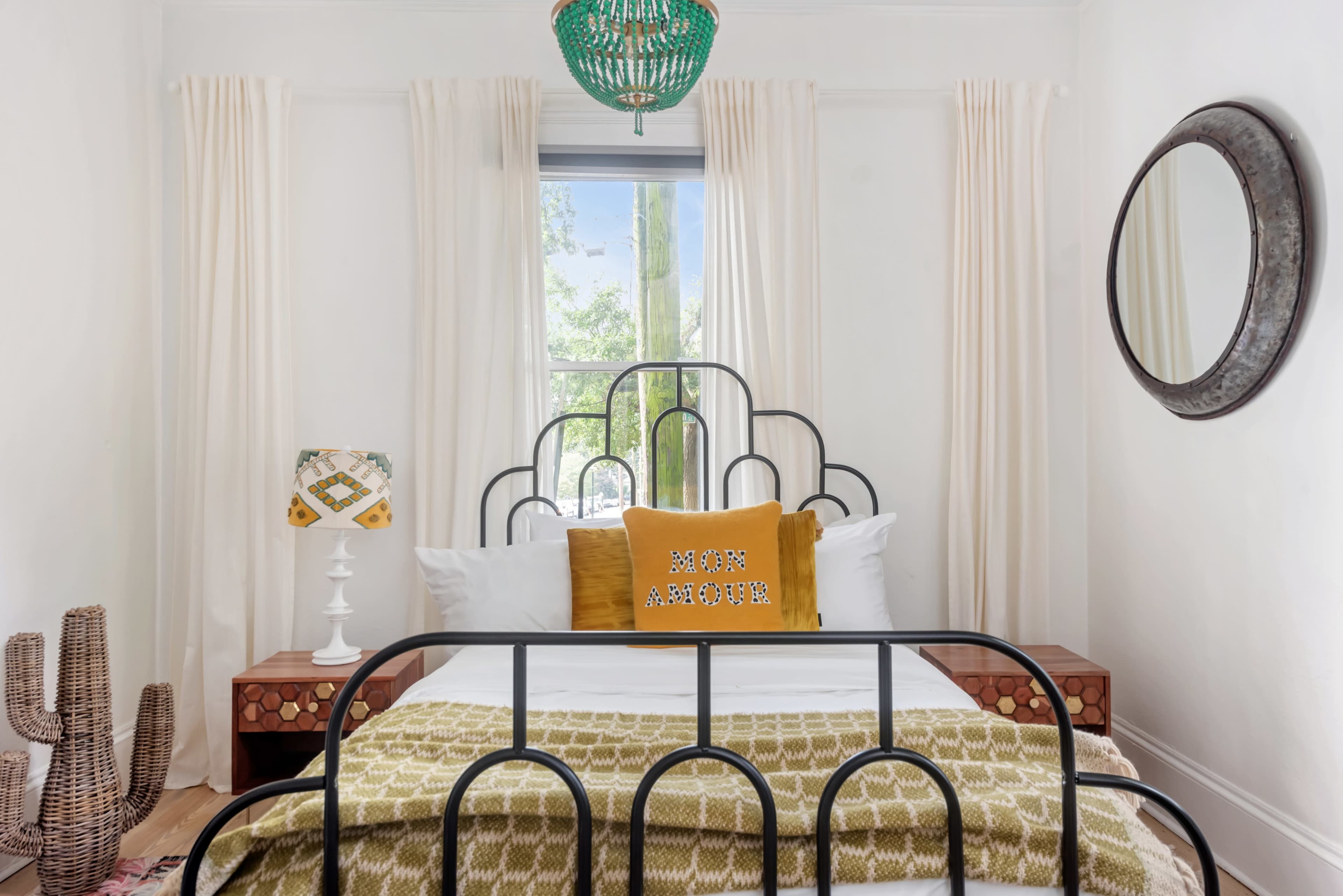 The image shows a cozy bedroom featuring a black metal bed frame with a patterned yellow and green blanket, a decorative pillow, a wooden nightstand with a lamp, a round mirror, and potted plants by the window.
