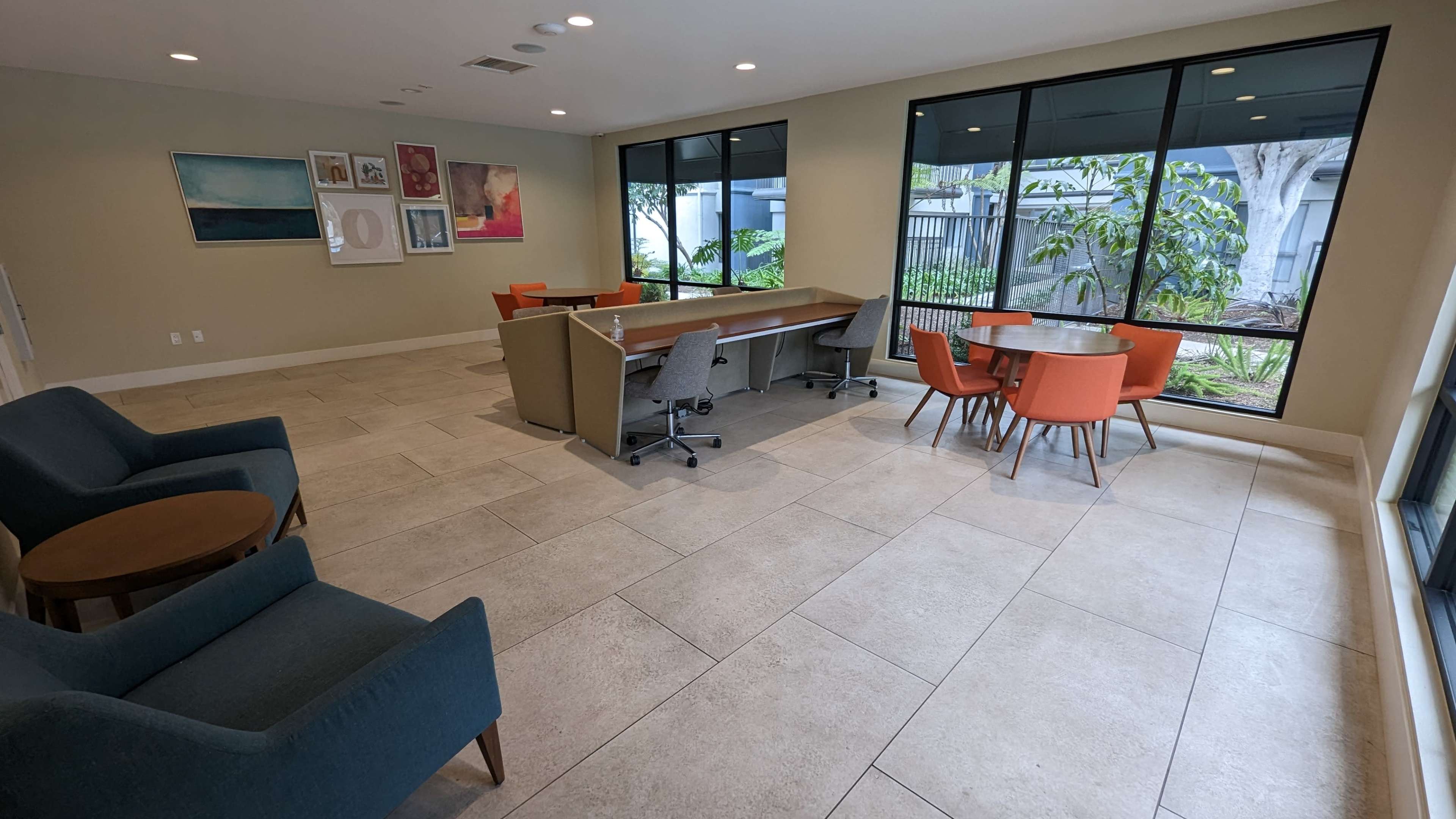 A modern communal lounge area with seating arrangements, a large table, and windows overlooking greenery.