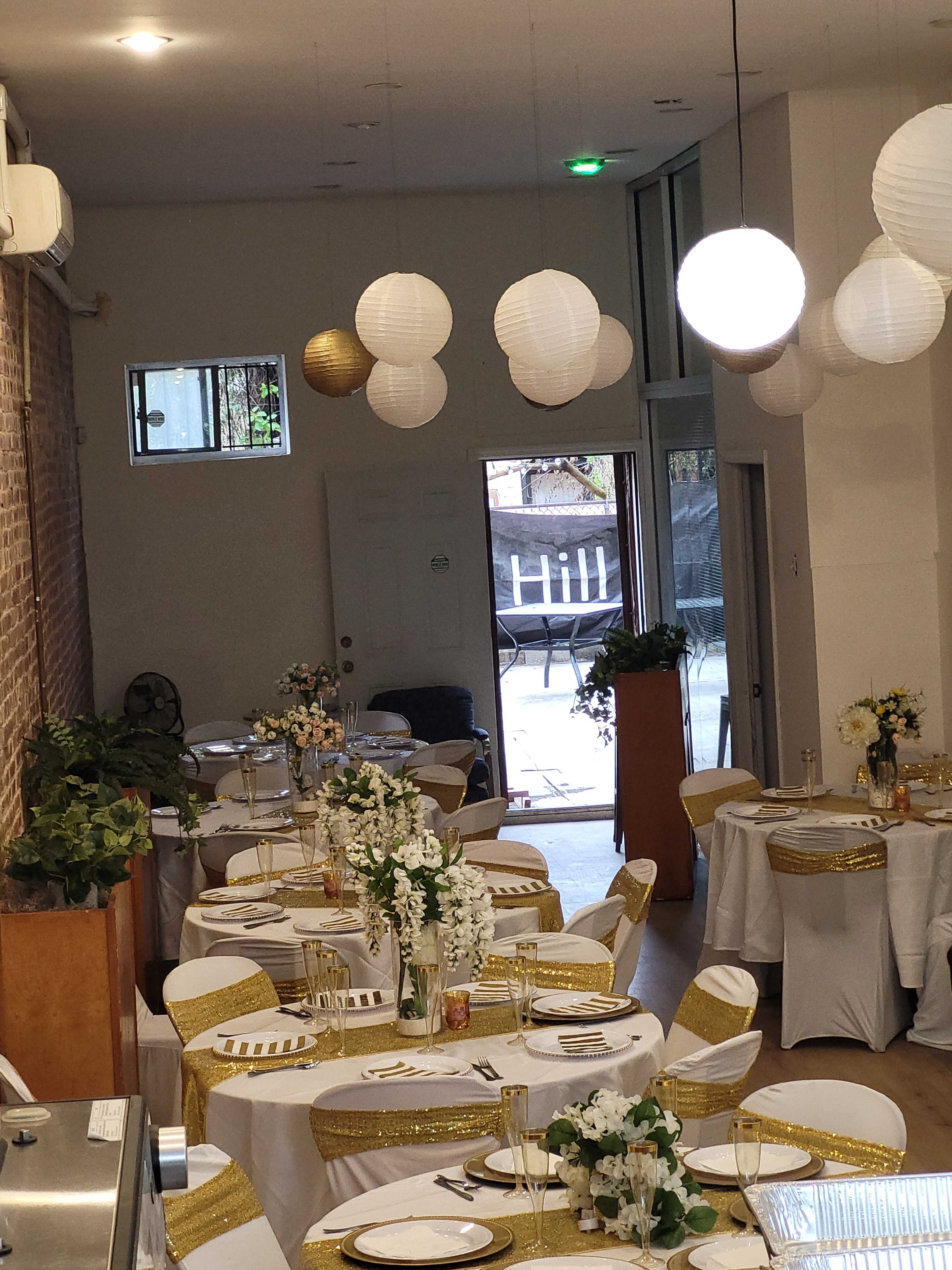 The image shows a banquet hall decorated with white tables adorned with gold accents and illuminated by hanging paper lanterns.