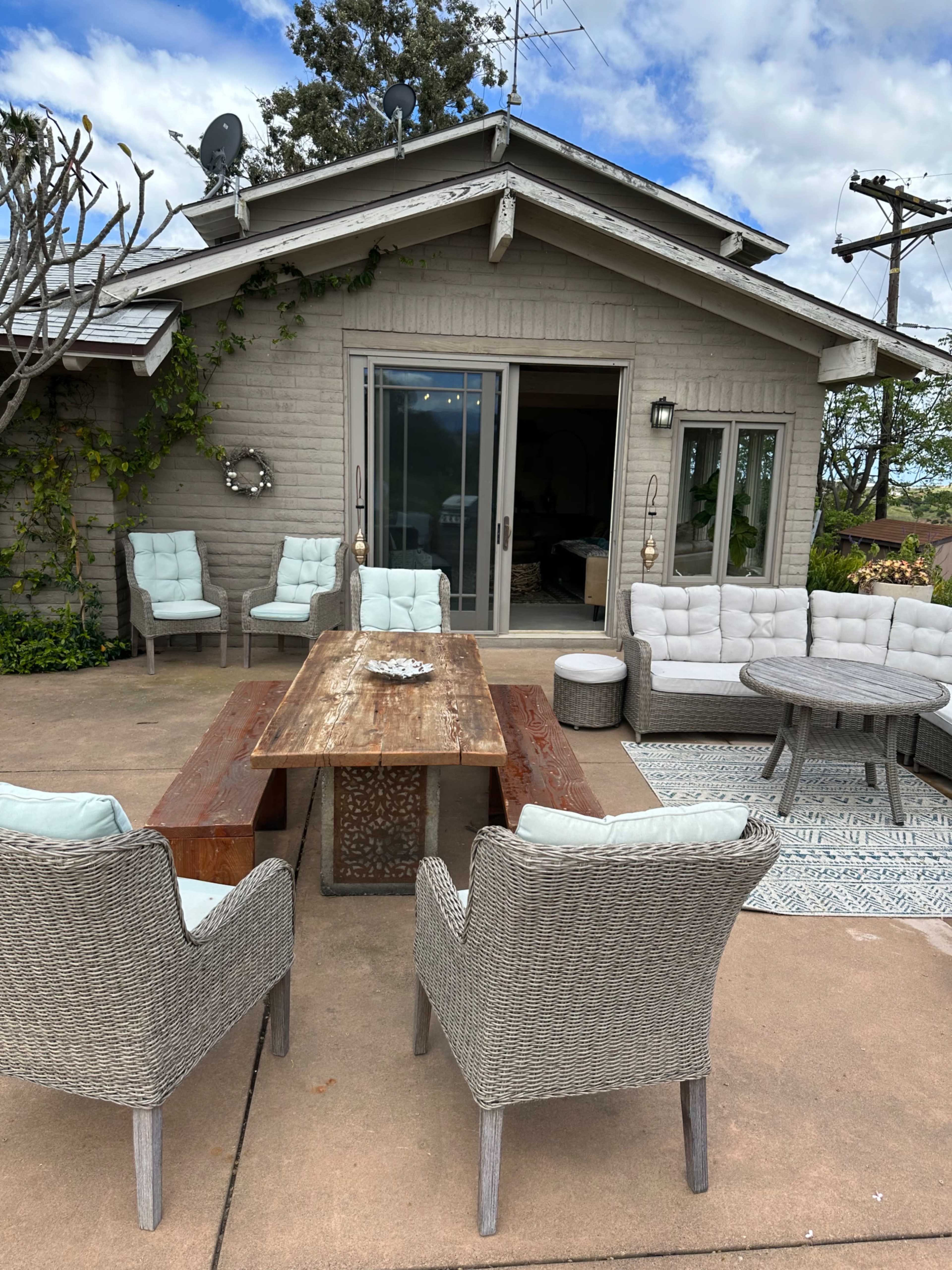 A patio area features a wooden table surrounded by various seating options and a view of a house with large sliding doors.