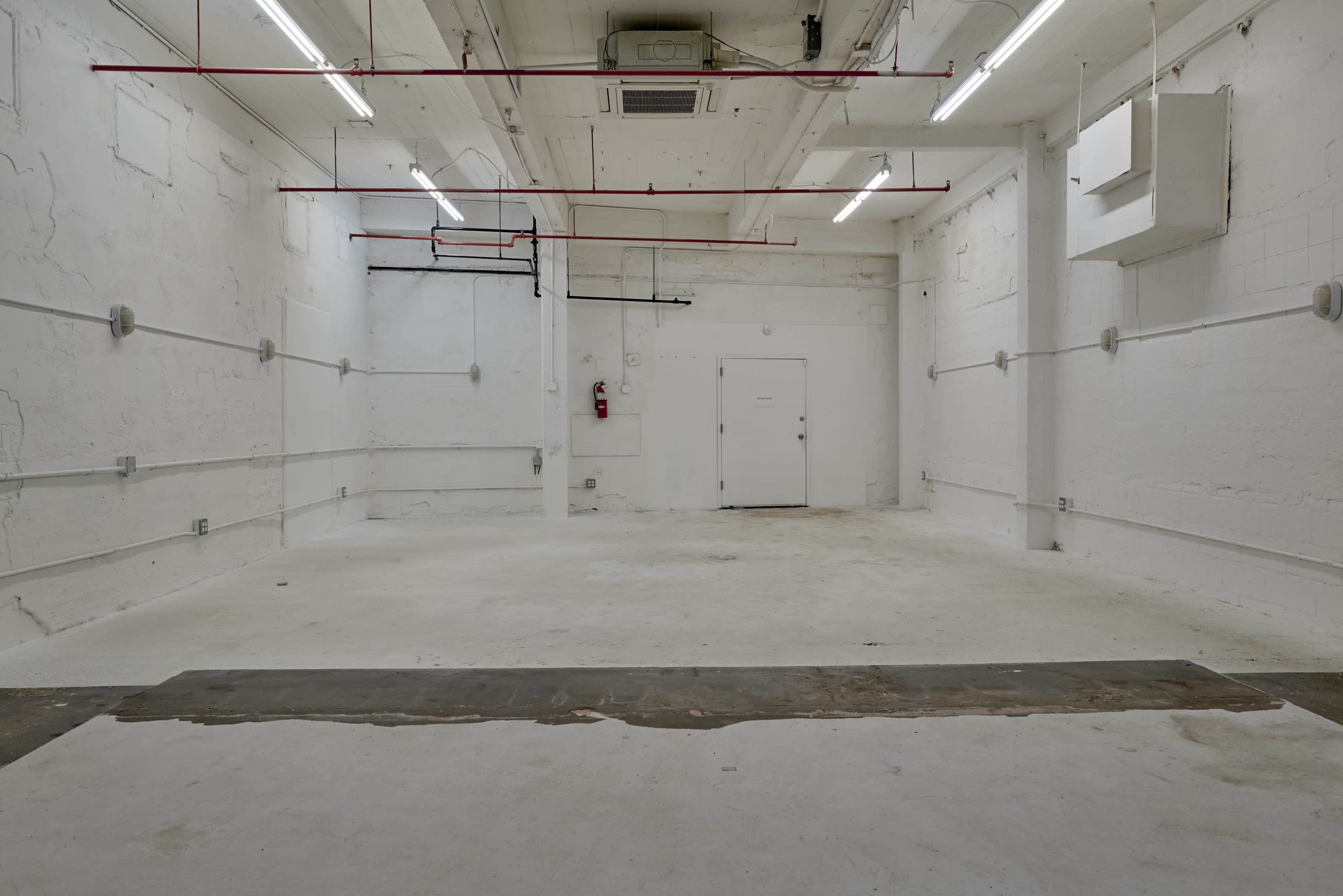 The image depicts a vacant, empty room with white walls, concrete flooring, and exposed overhead fixtures.