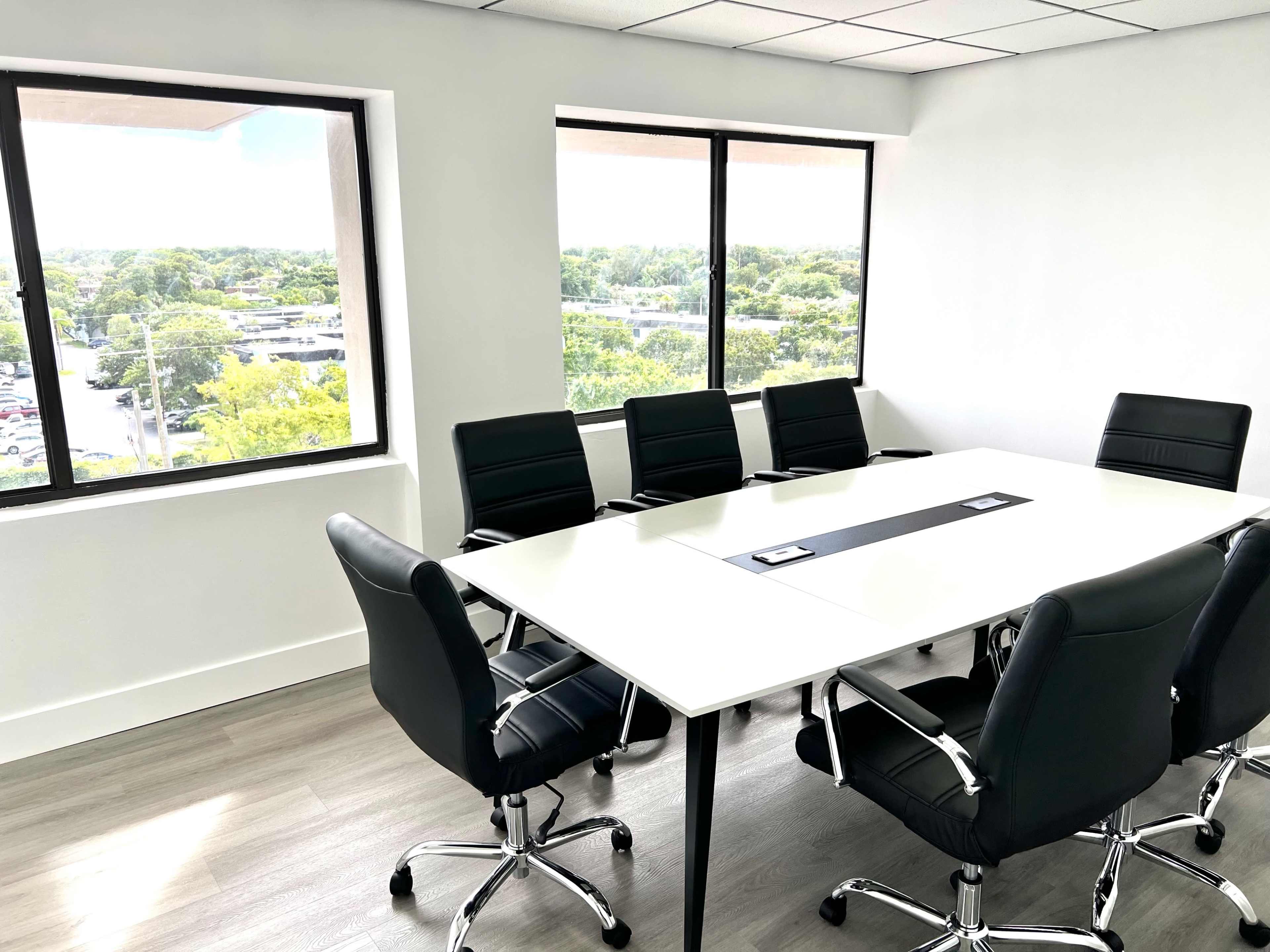 A modern conference room features a large white table surrounded by black leather chairs, with windows offering a view of greenery and a parking lot outside.
