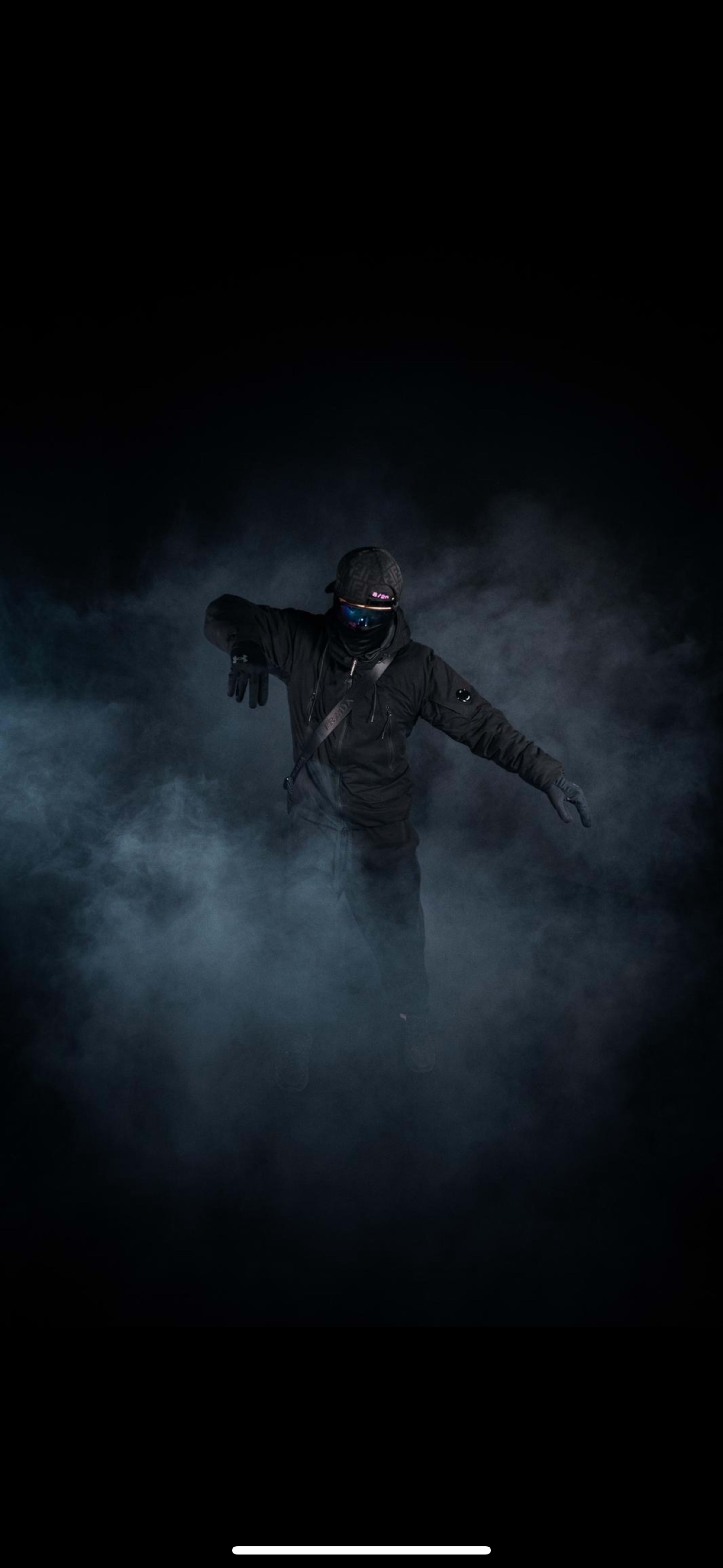 A figure dressed in dark clothing and gloves is posed amidst a dense, smoky background.