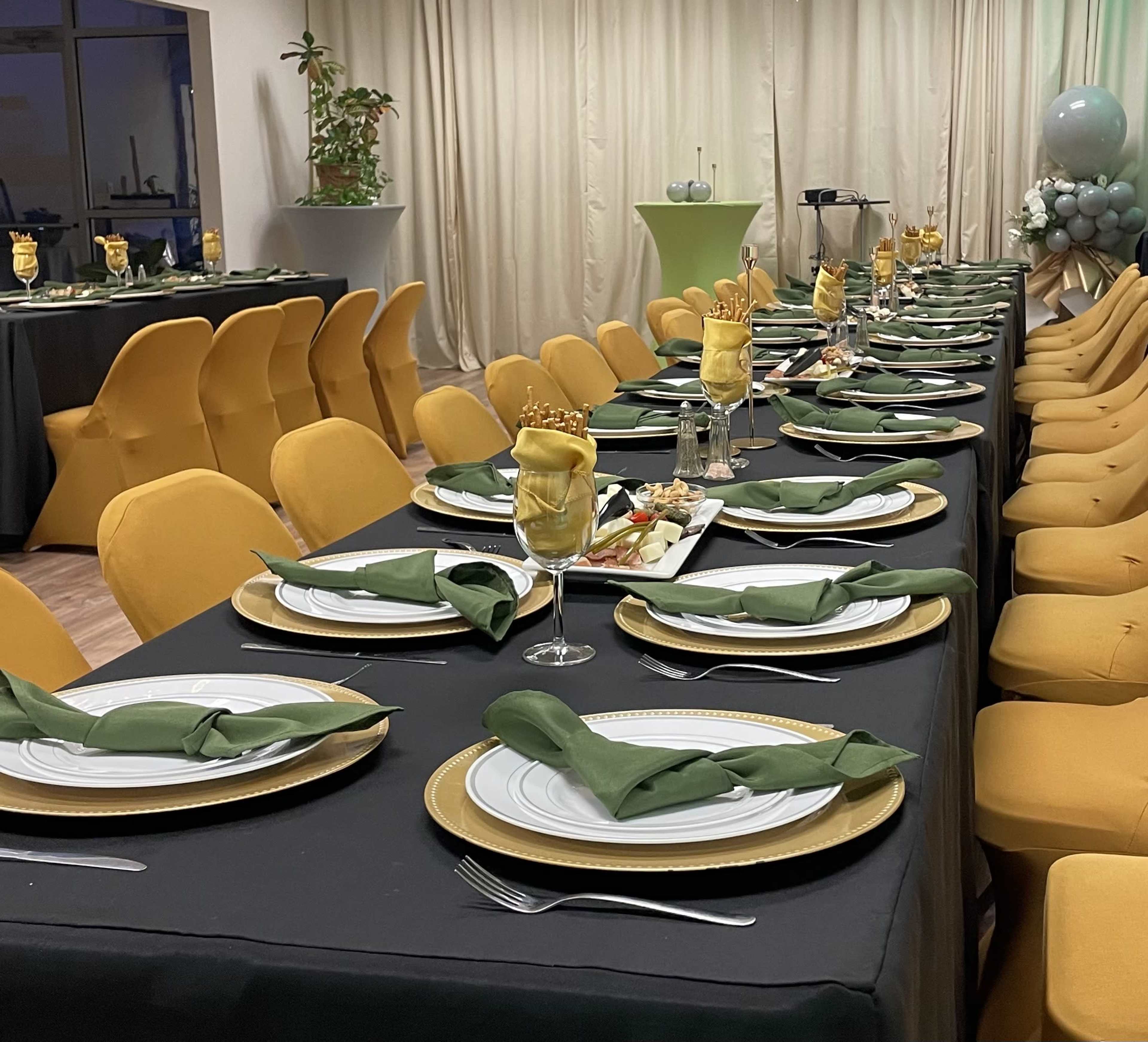A long dining table is set with green napkins and gold plates, surrounded by yellow chairs in a decorated event space.