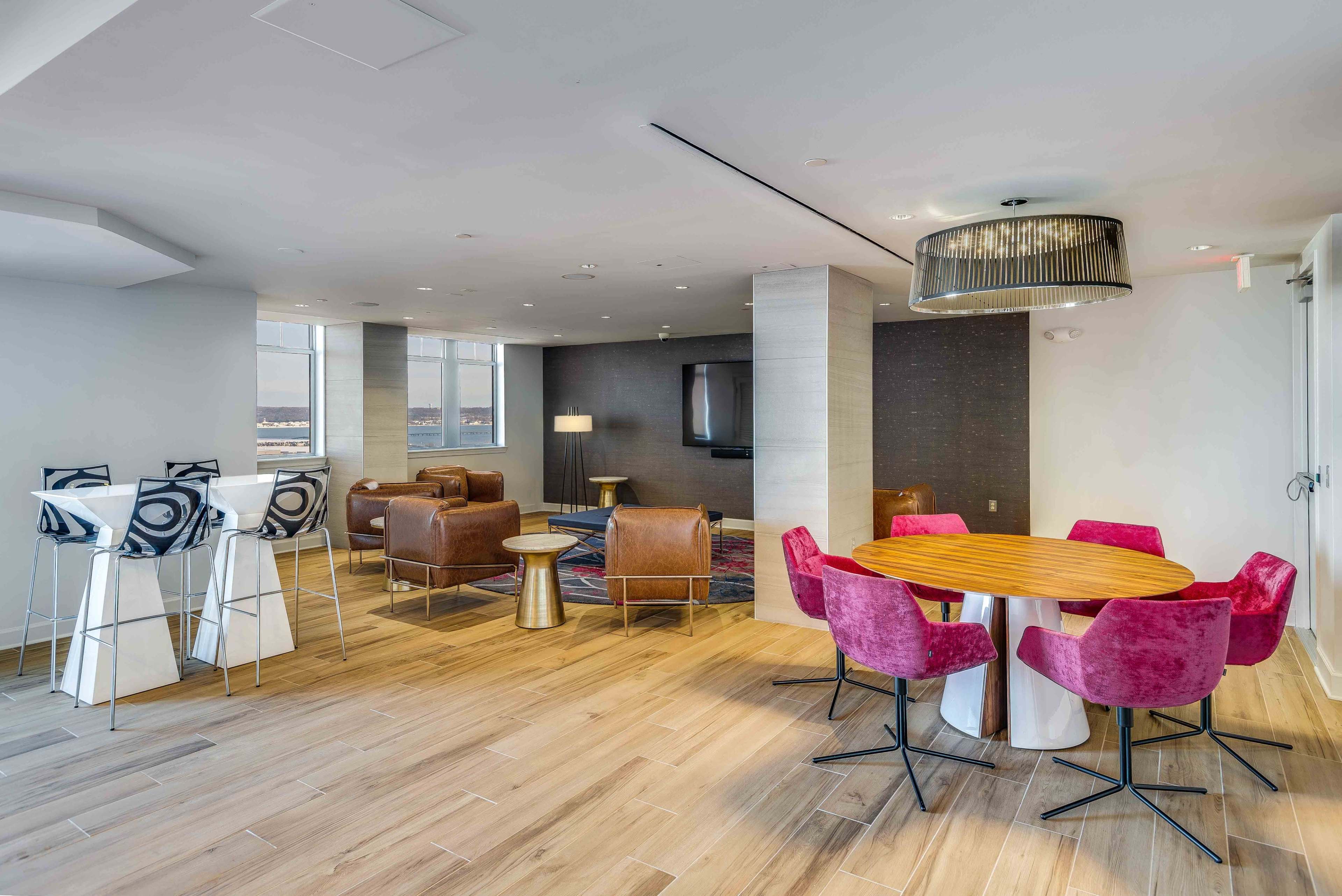 A modern, open-concept lounge area featuring a circular dining table surrounded by pink chairs, a seating arrangement with brown leather couches, and large windows providing a view.