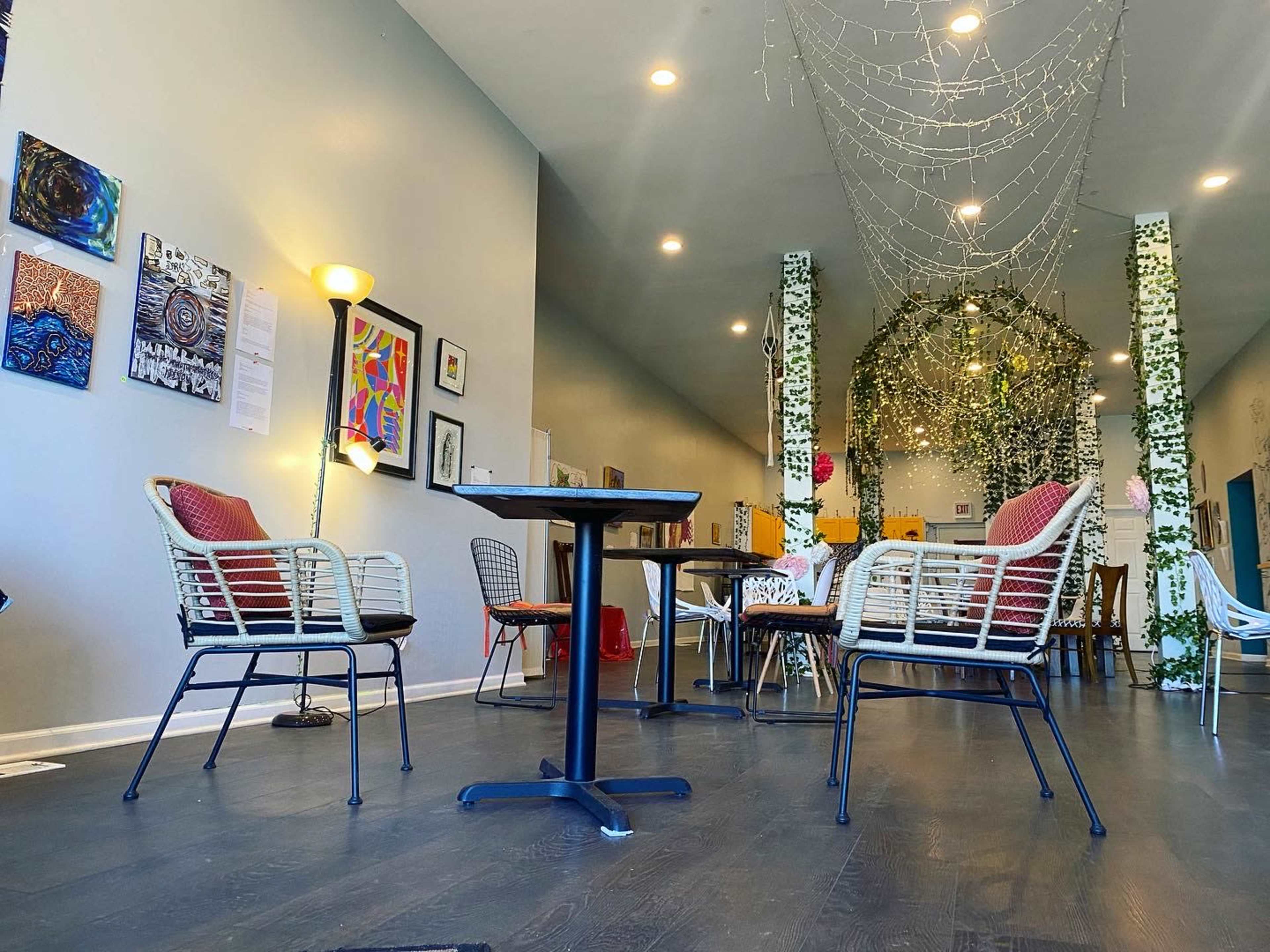 A spacious, well-lit cafe featuring several round tables and chairs, with art on the walls and decorative greenery overhead.