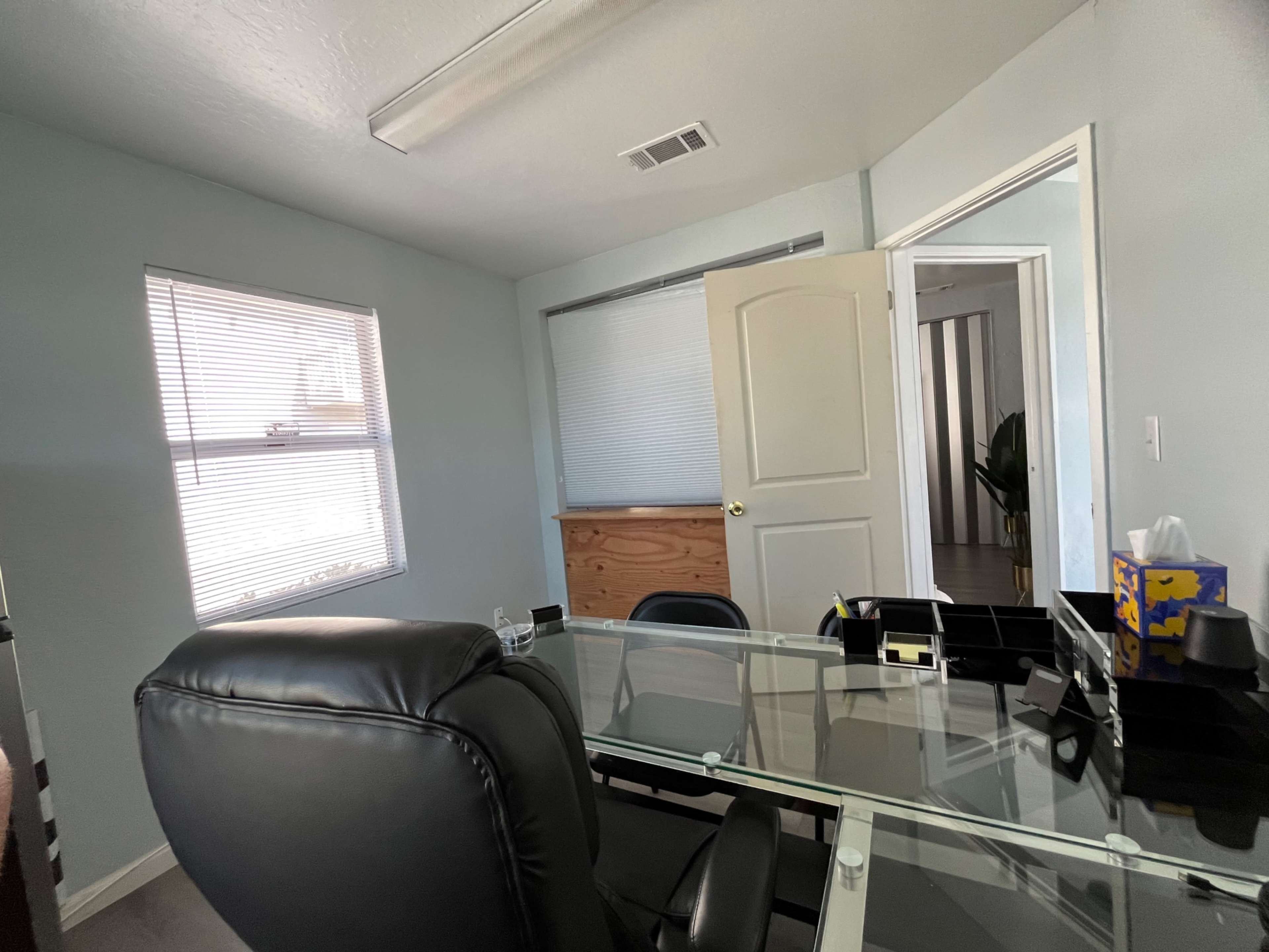 The image shows a small office with a glass desk, black office chairs, and a window, along with an open door leading to another room.