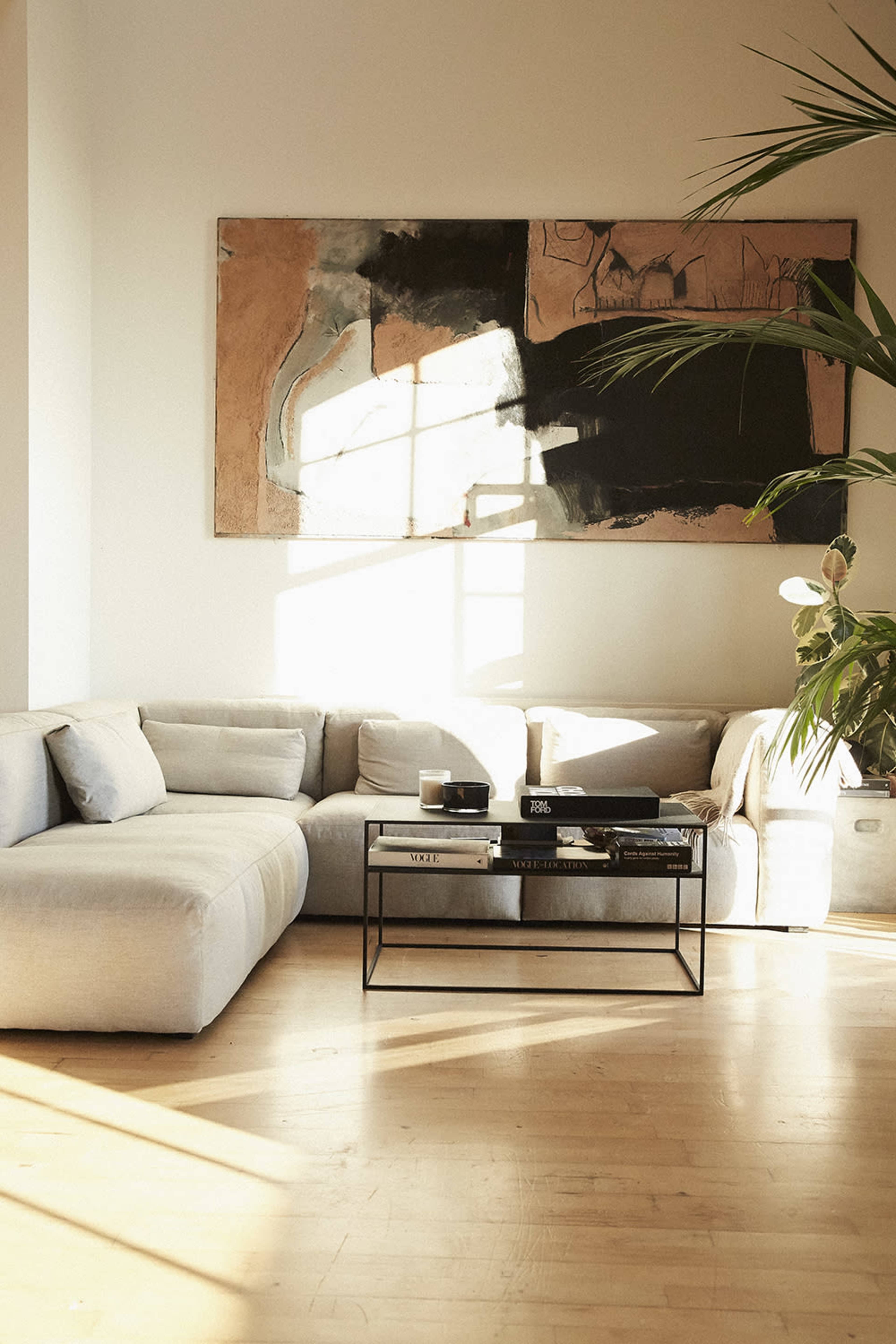 A bright living room features a light-colored sectional sofa, a black coffee table with various items on it, and a large abstract painting on the wall, illuminated by natural sunlight.