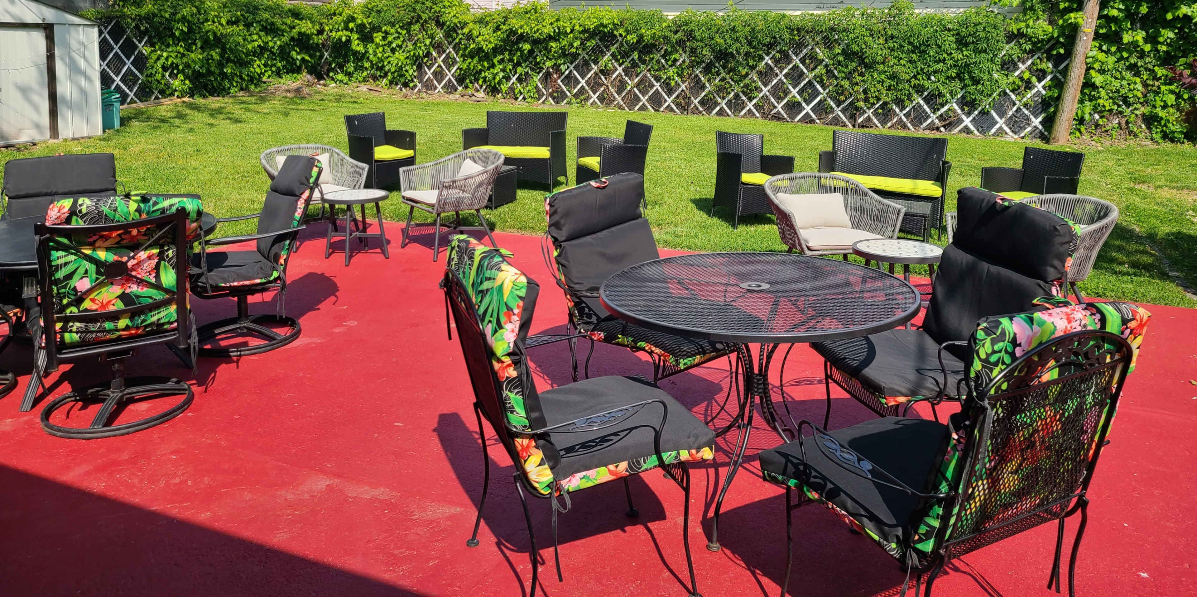 A patio area with various styles of outdoor chairs and tables arranged on a red surface, surrounded by green grass and a vine-covered fence.