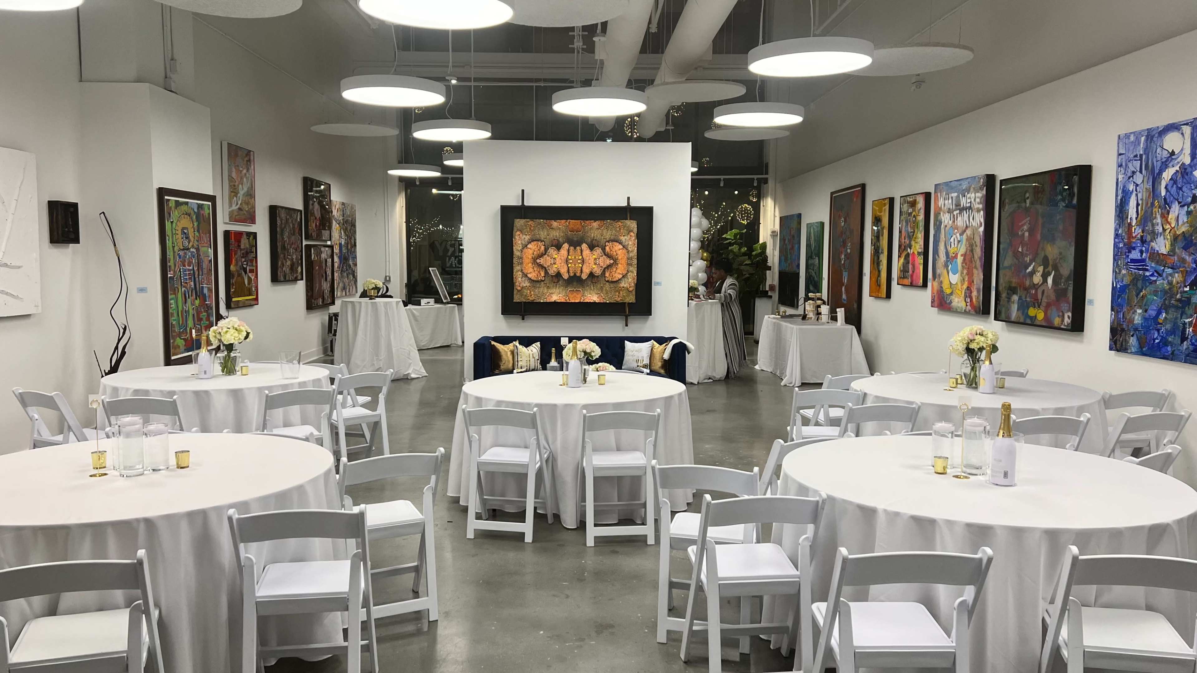 A gallery-style room set up for an event, featuring several round tables covered with white tablecloths, surrounded by artwork hanging on the walls.