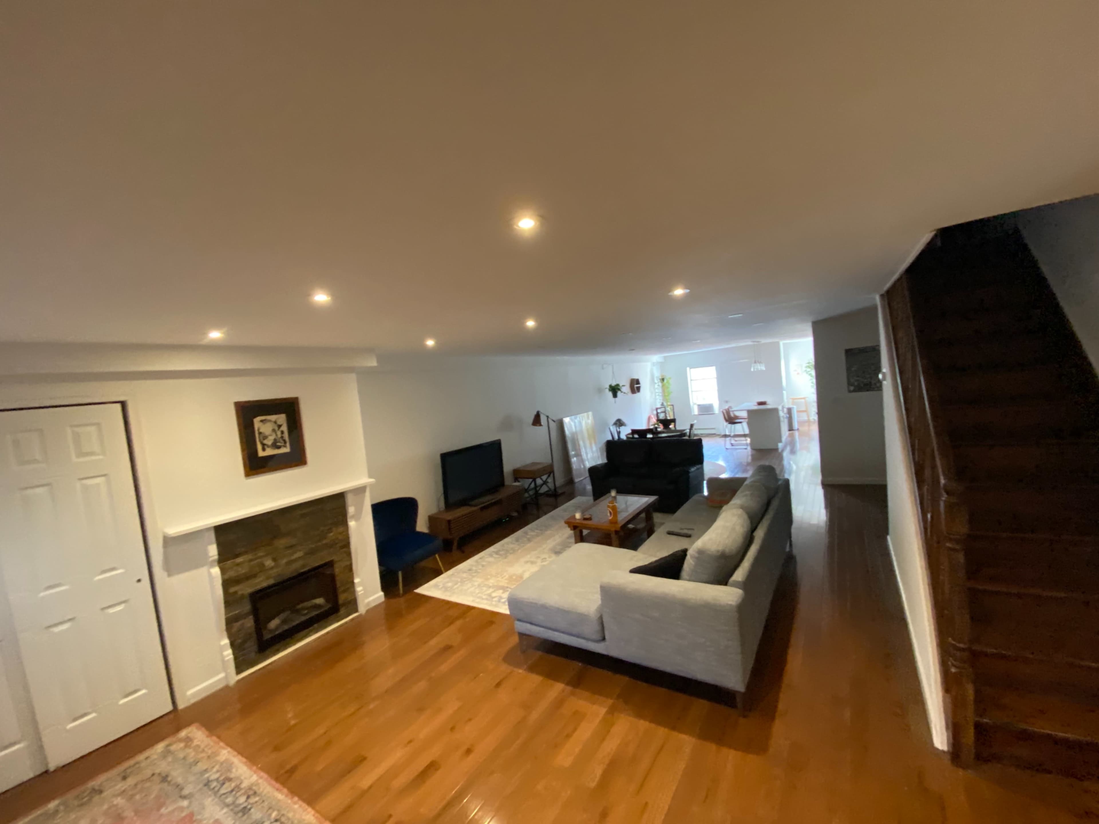 A living room with hardwood flooring, a stone fireplace, a gray sectional sofa, a television, and a staircase in the corner.