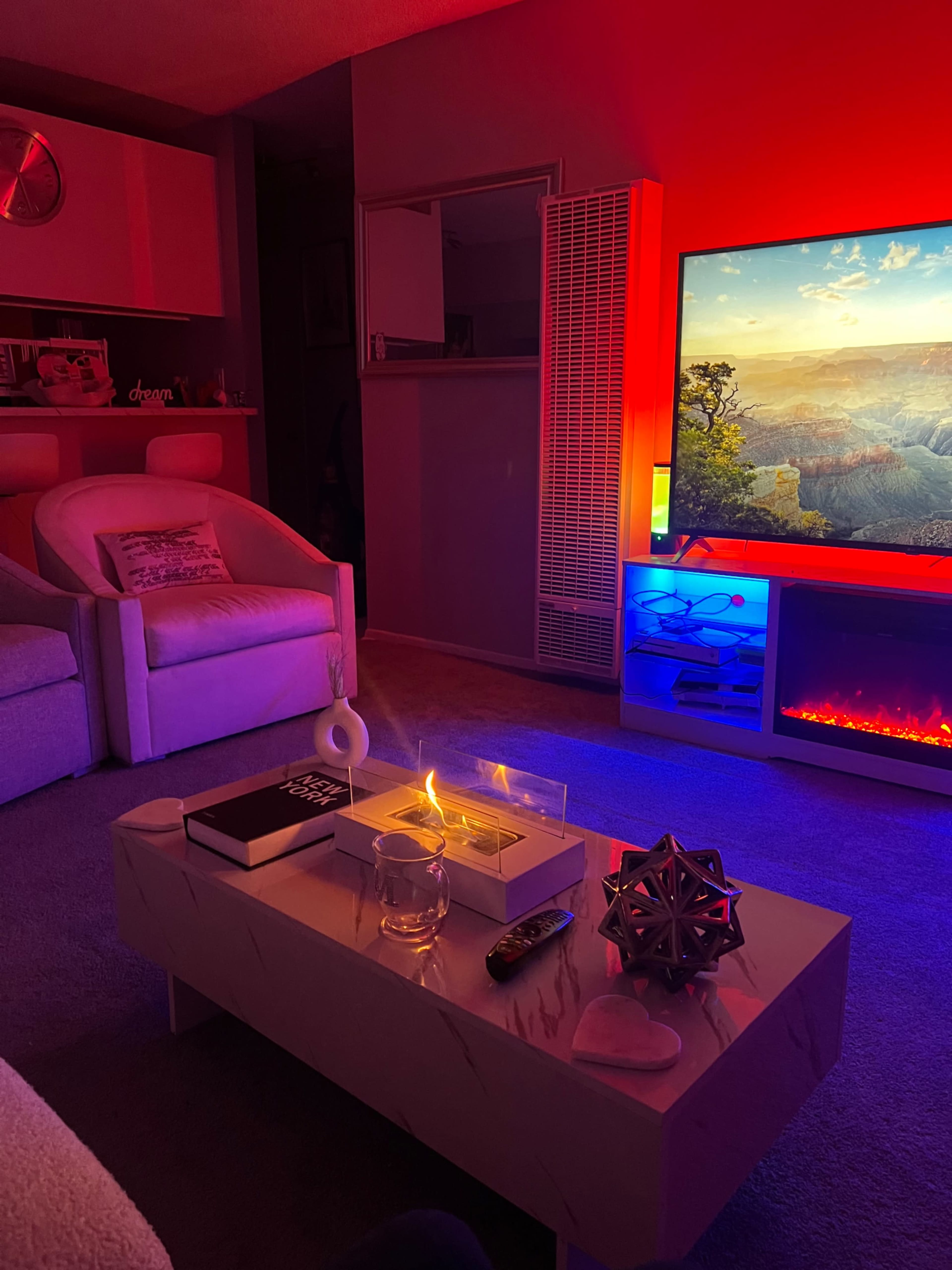 A modern living room features a cozy seating area with a light-colored armchair and a coffee table adorned with decorative objects, illuminated by soft, colorful lighting from the television and wall fixtures.