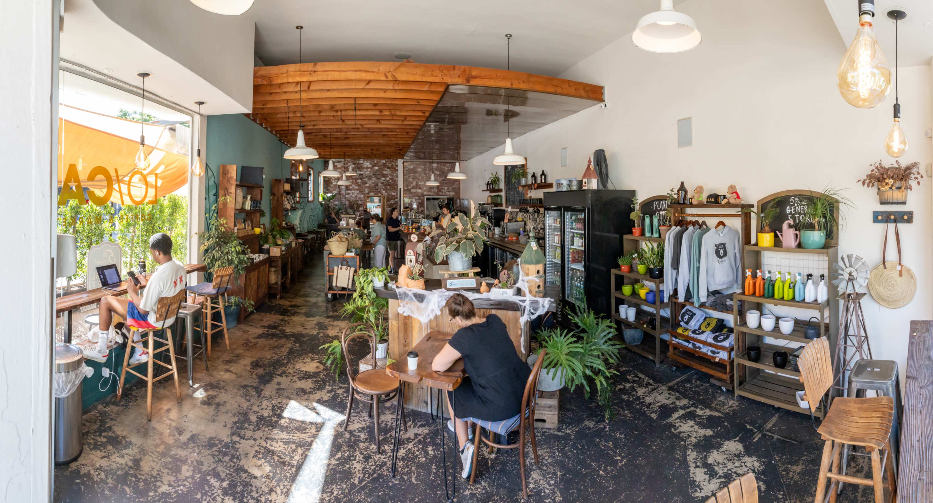 A bright and spacious café with wooden beams, plants, and various seating areas where patrons are working and enjoying their time.