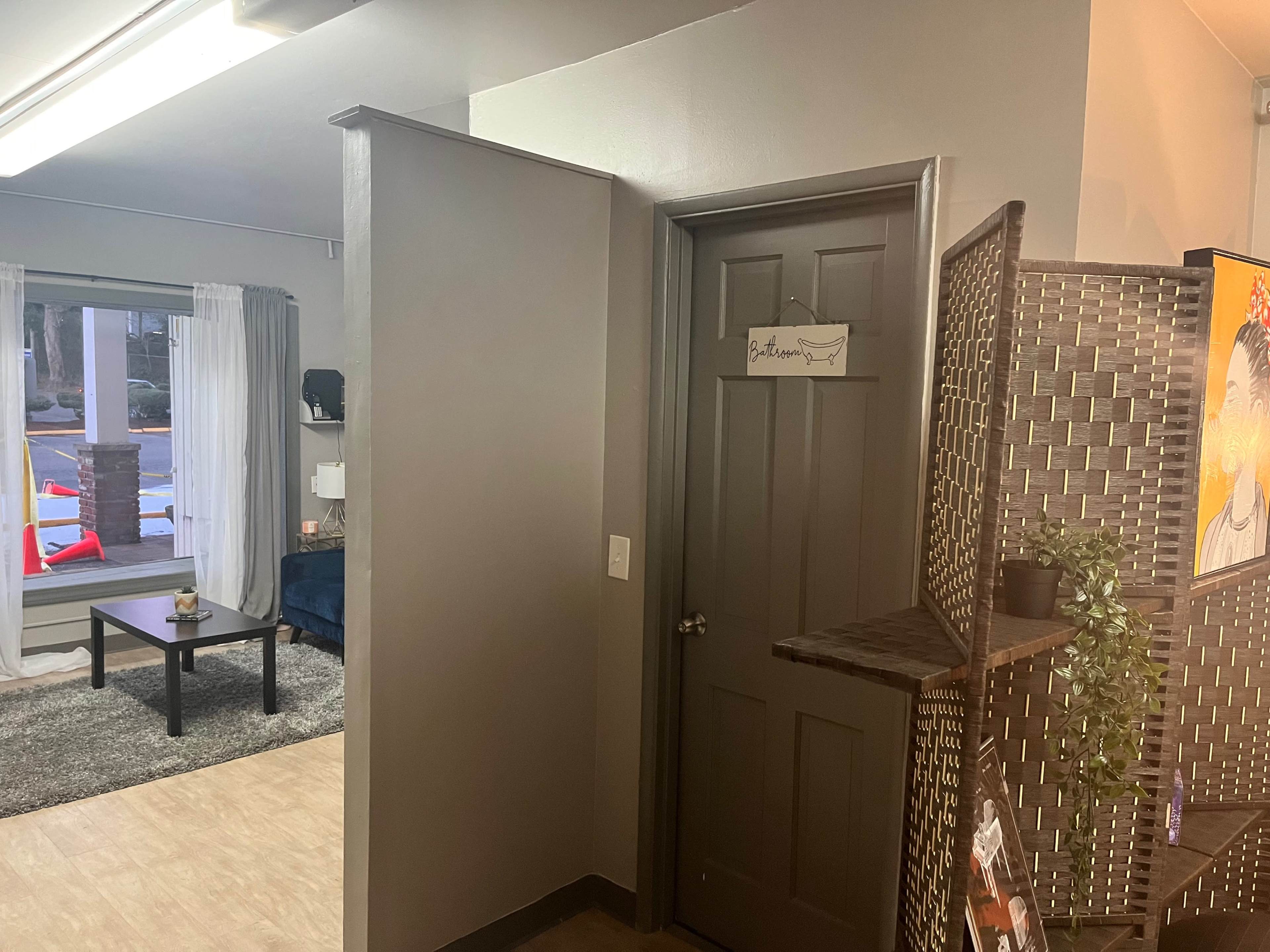 A corner of a room with a doorway labeled "Bathroom" and a view into a living space featuring a table and couch through a window.