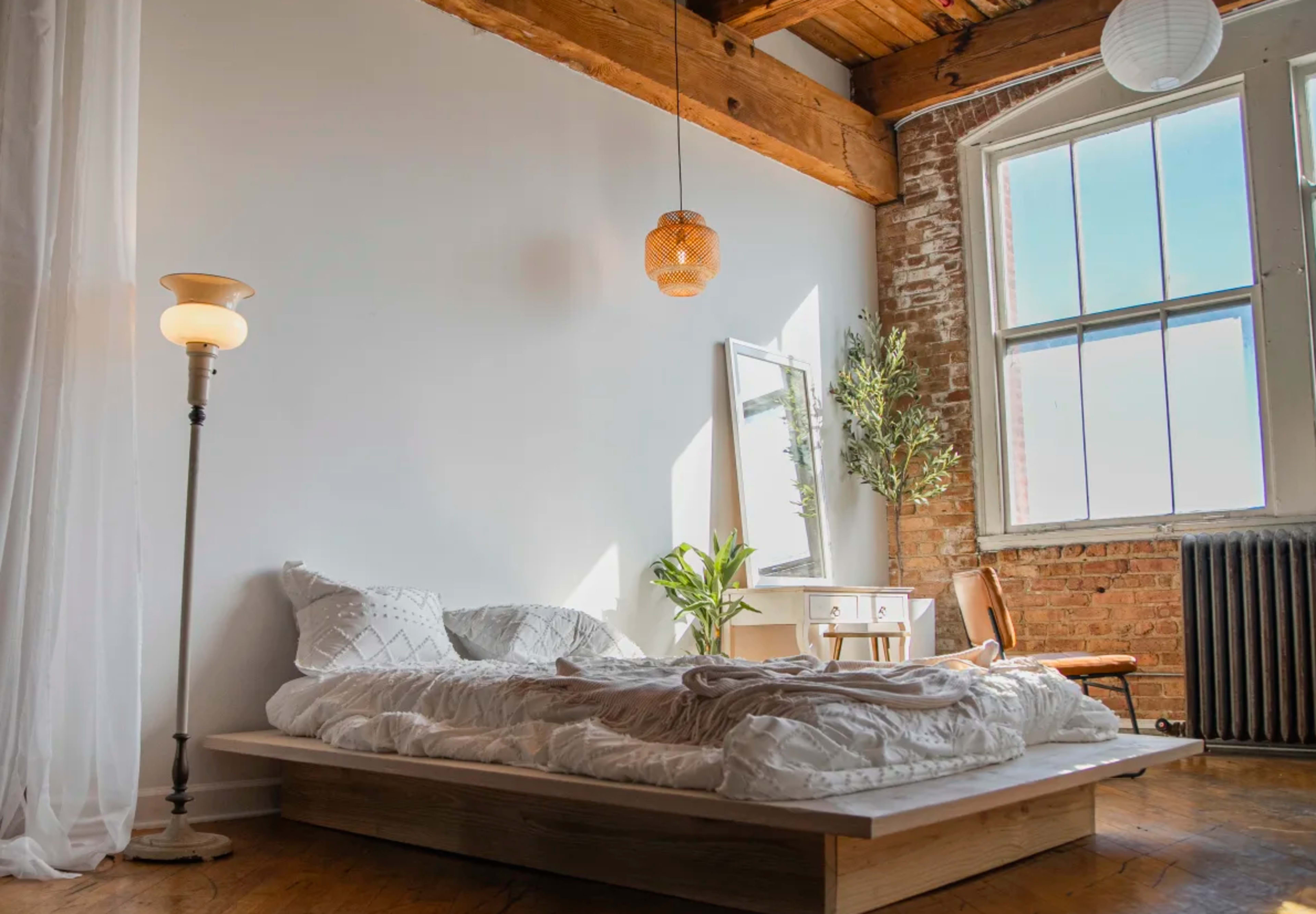 The image shows a minimalist bedroom with a low wooden bed, natural light streaming through a large window, and a small desk accompanied by a chair and decorative plants.