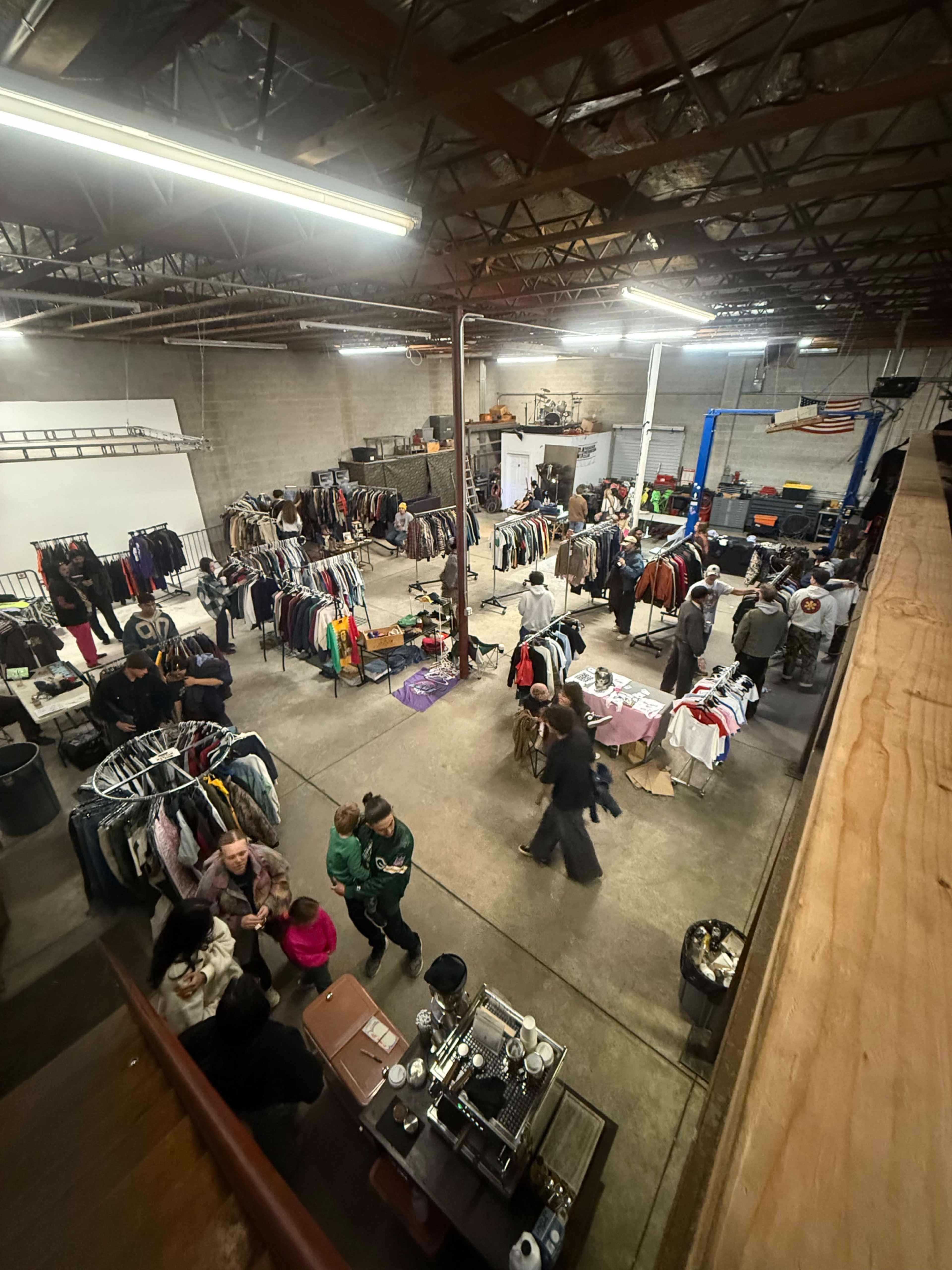 The image shows an indoor clothing sale event in a warehouse, featuring racks of clothes and various groups of people browsing and socializing.