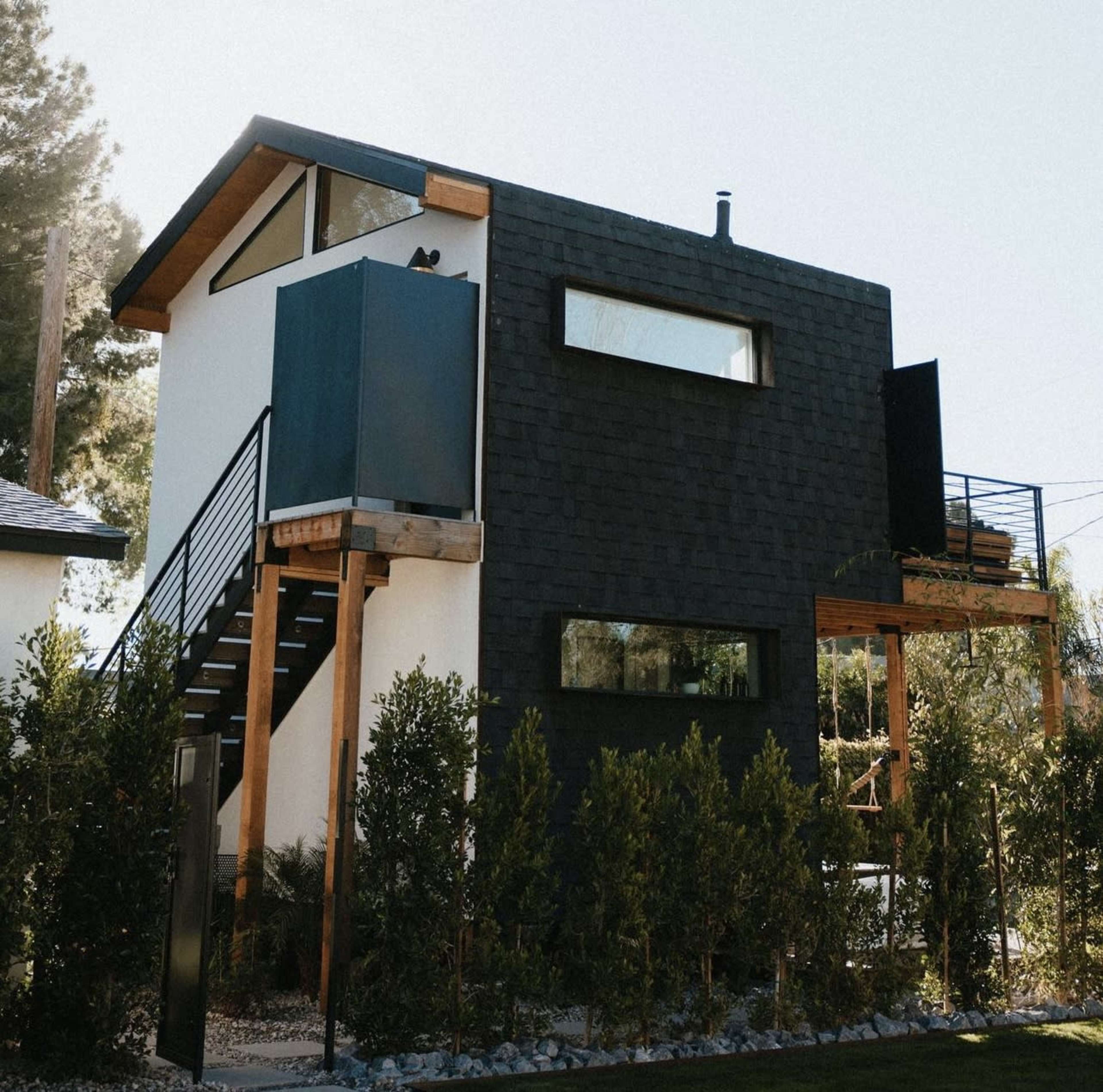 The image shows a modern two-story house with a black exterior and wooden accents, surrounded by neatly trimmed shrubs.