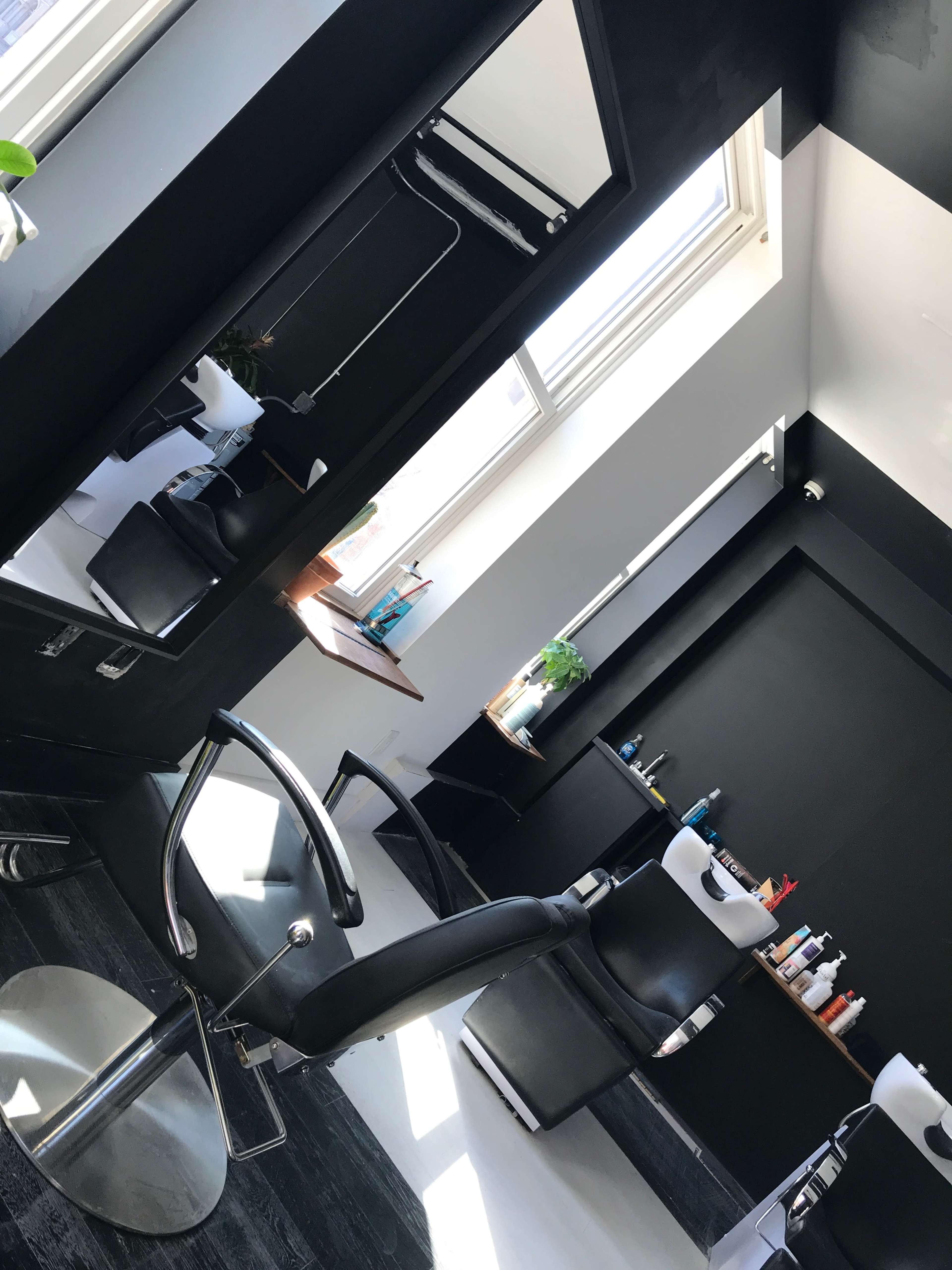 A modern hair salon with black walls, two salon chairs, and large mirrors, illuminated by natural light from a window.