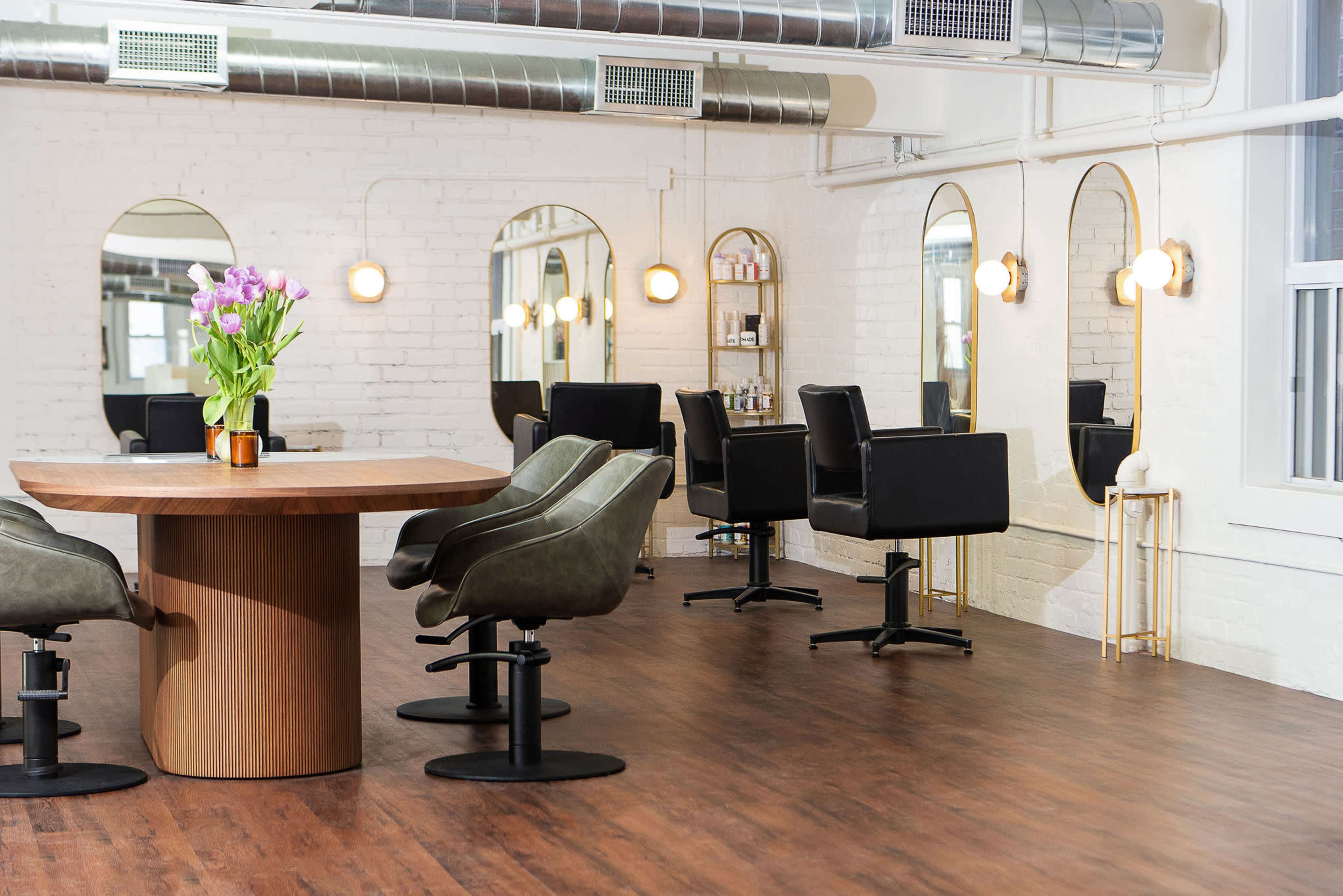 A modern hair salon featuring a round wooden table with a floral arrangement, black salon chairs, and large mirrors along the walls.