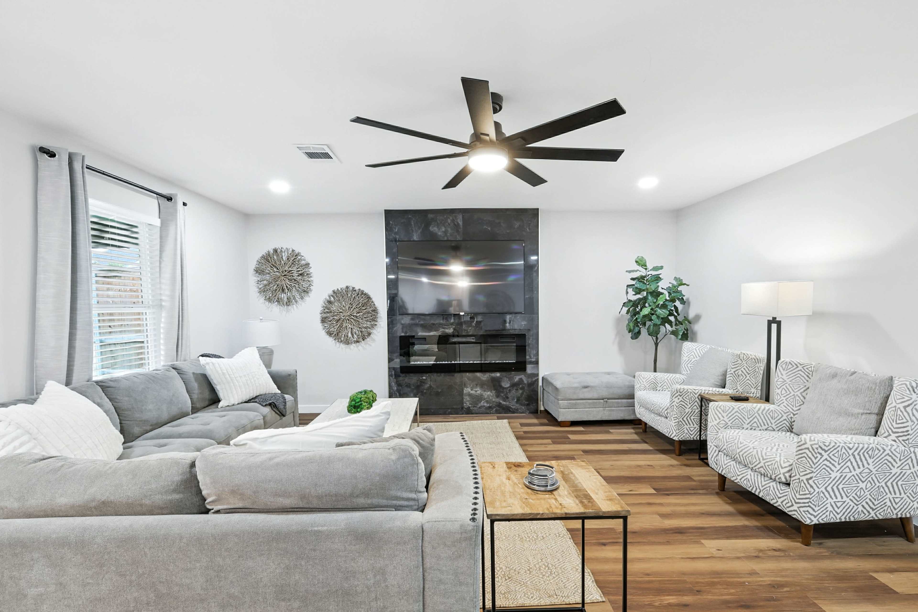 The image features a modern living room with a grey sectional sofa, two patterned armchairs, a wooden coffee table, and a wall-mounted television above a black accent wall.