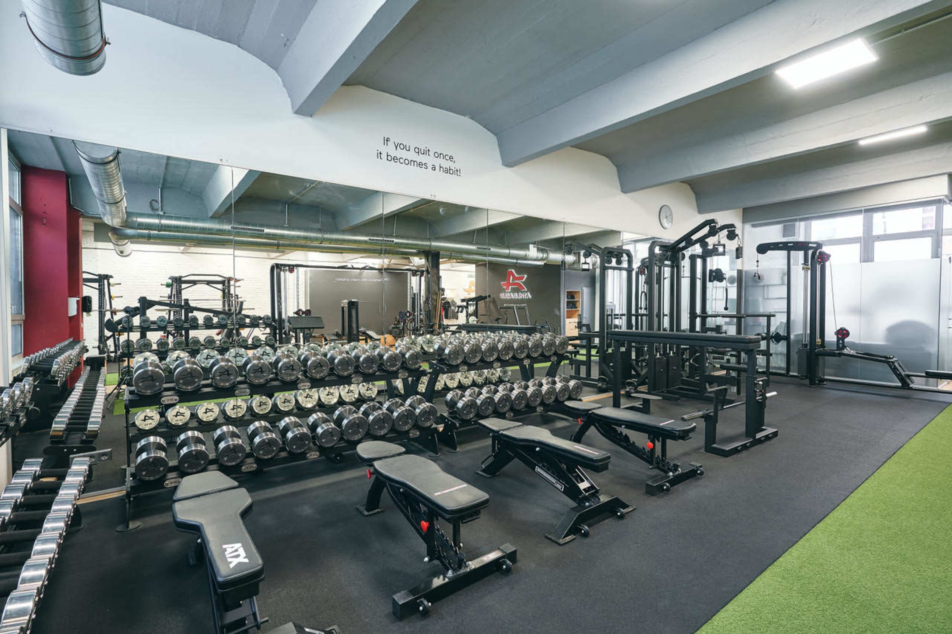 The image shows a well-equipped gym featuring a row of free weights and multiple weight training machines arranged in a spacious area with rubber flooring.