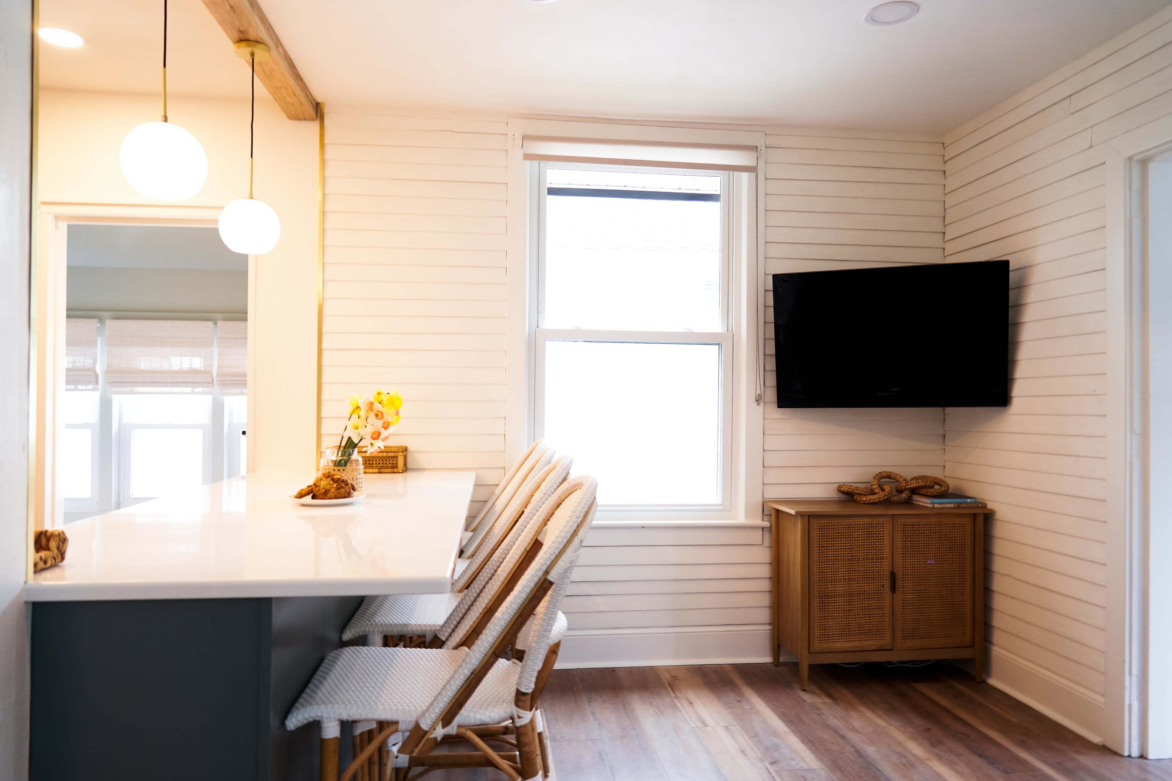 A bright kitchen area features a counter with four rattan chairs, a wall-mounted television, and large windows.